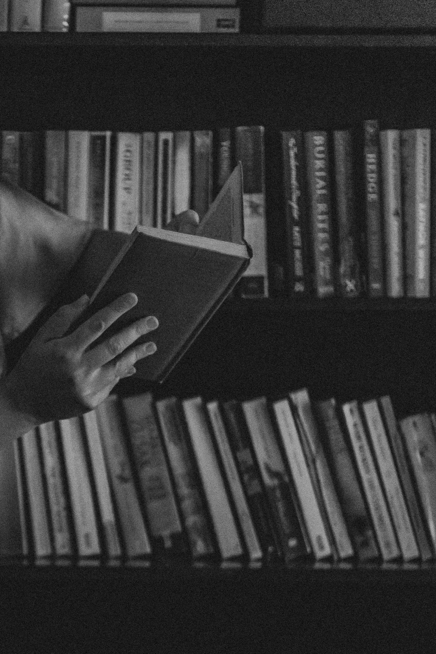 A hand reaches for a book on a shelf filled with various hardcover volumes in a moody black and white photograph.