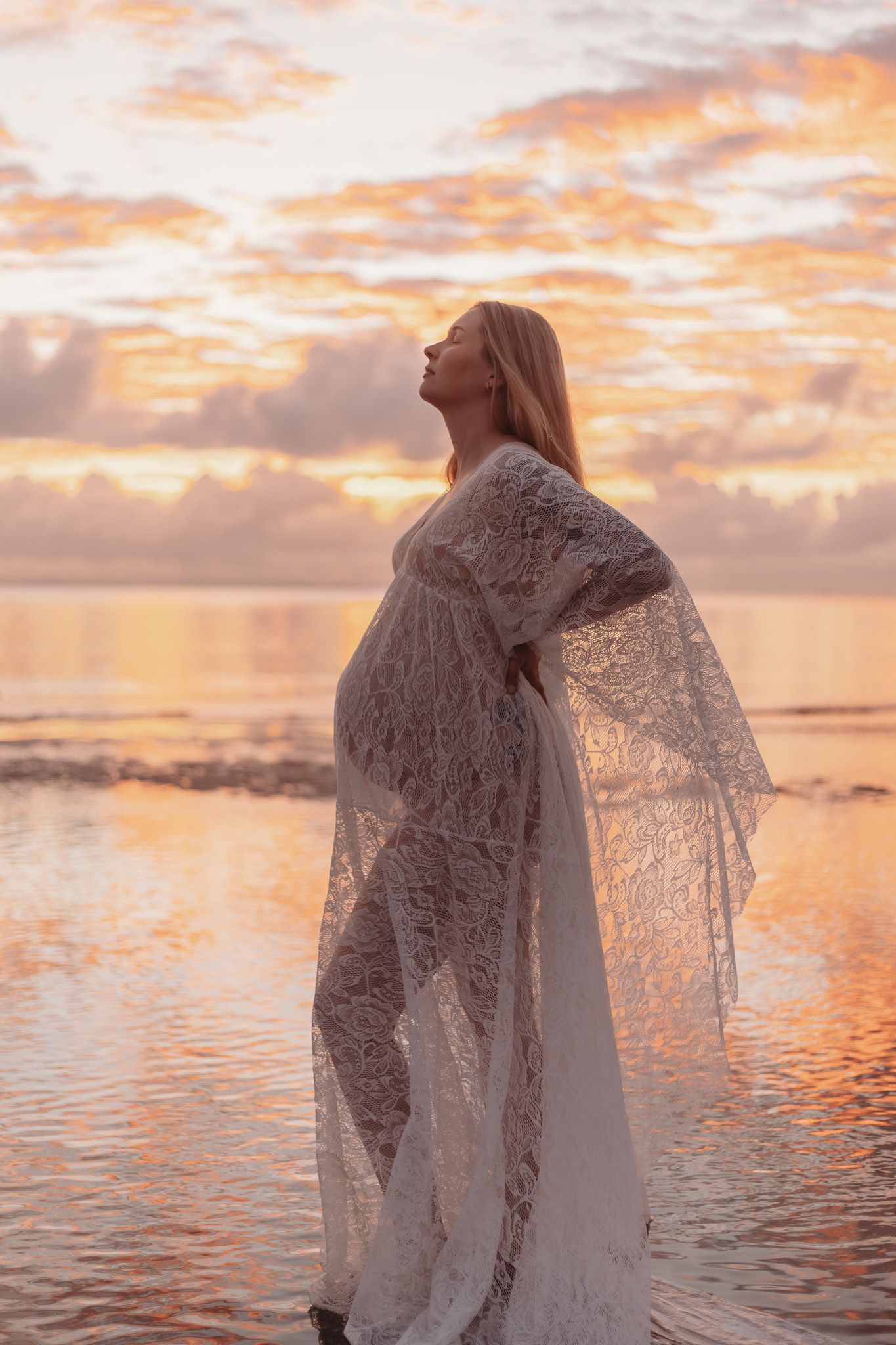 A figure in a flowing lace dress stands at the ocean's edge during a golden sunset with dramatic clouds in the sky.