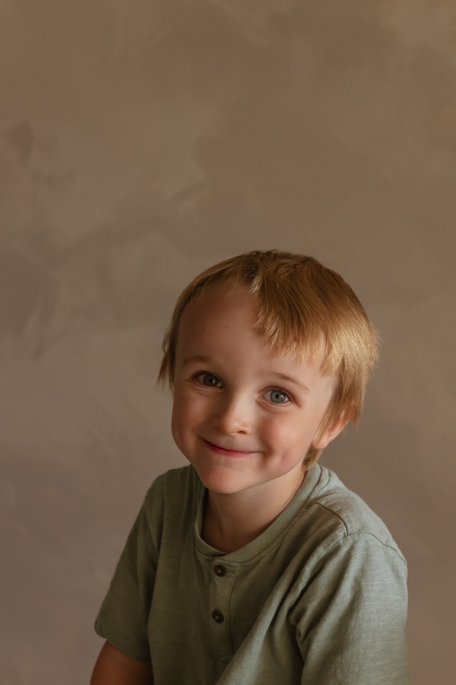 A young child wearing a sage green henley shirt poses against a tan background with a gentle expression.