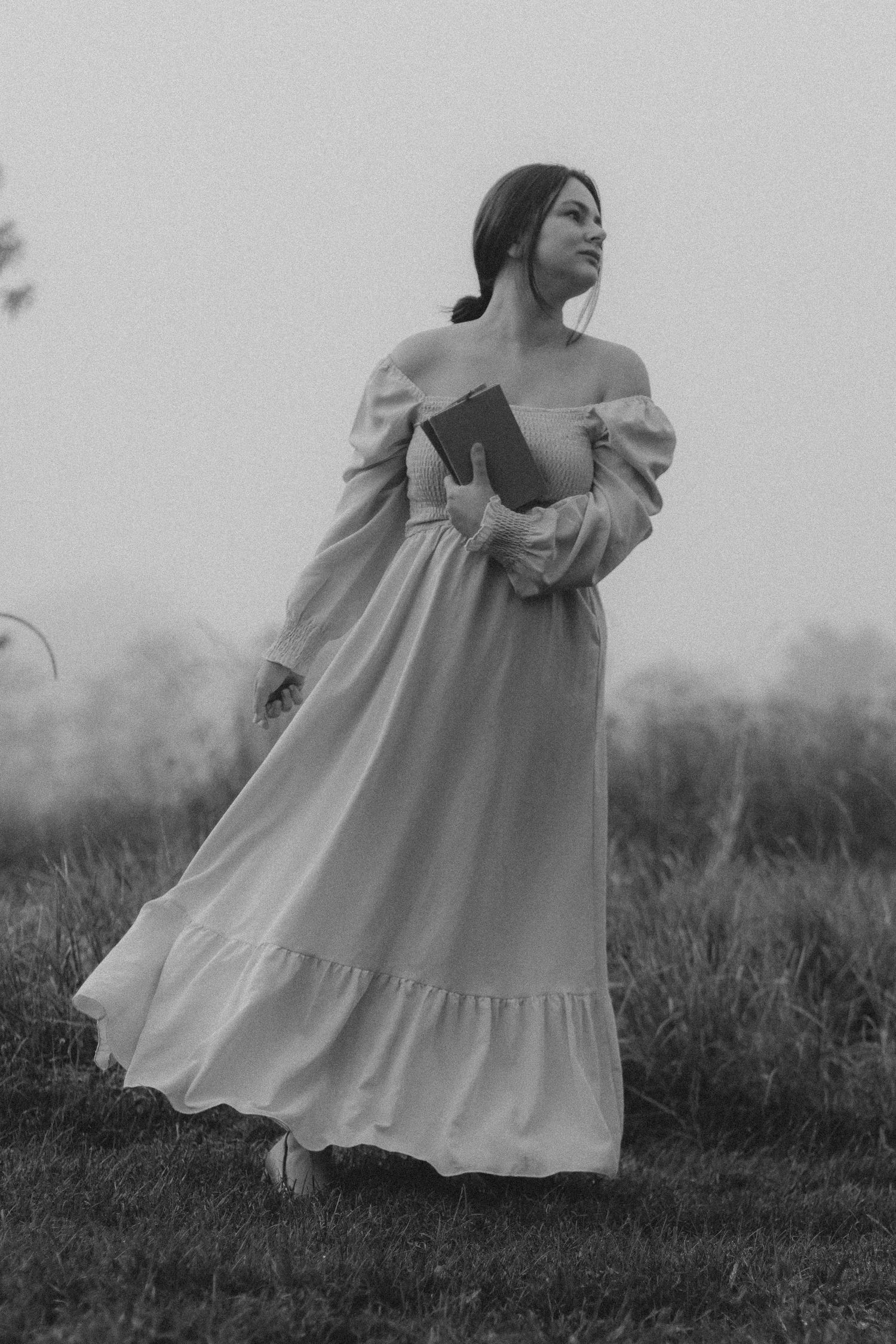 Black and white portrait of woman in flowing off-shoulder vintage dress standing in misty field at dusk.