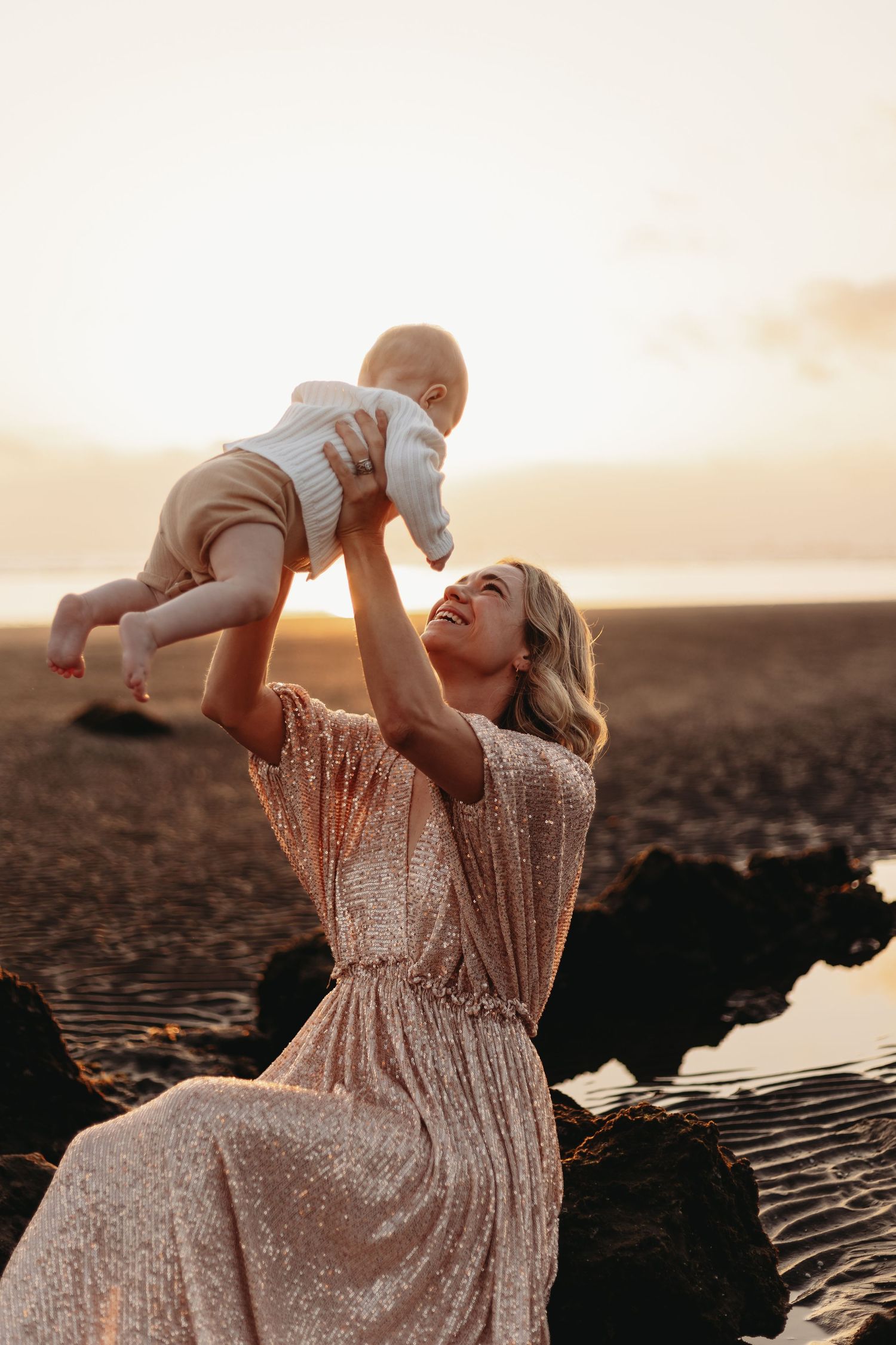 A parent lifts a baby joyfully against a golden sunset on a beach with volcanic rocks in the background.