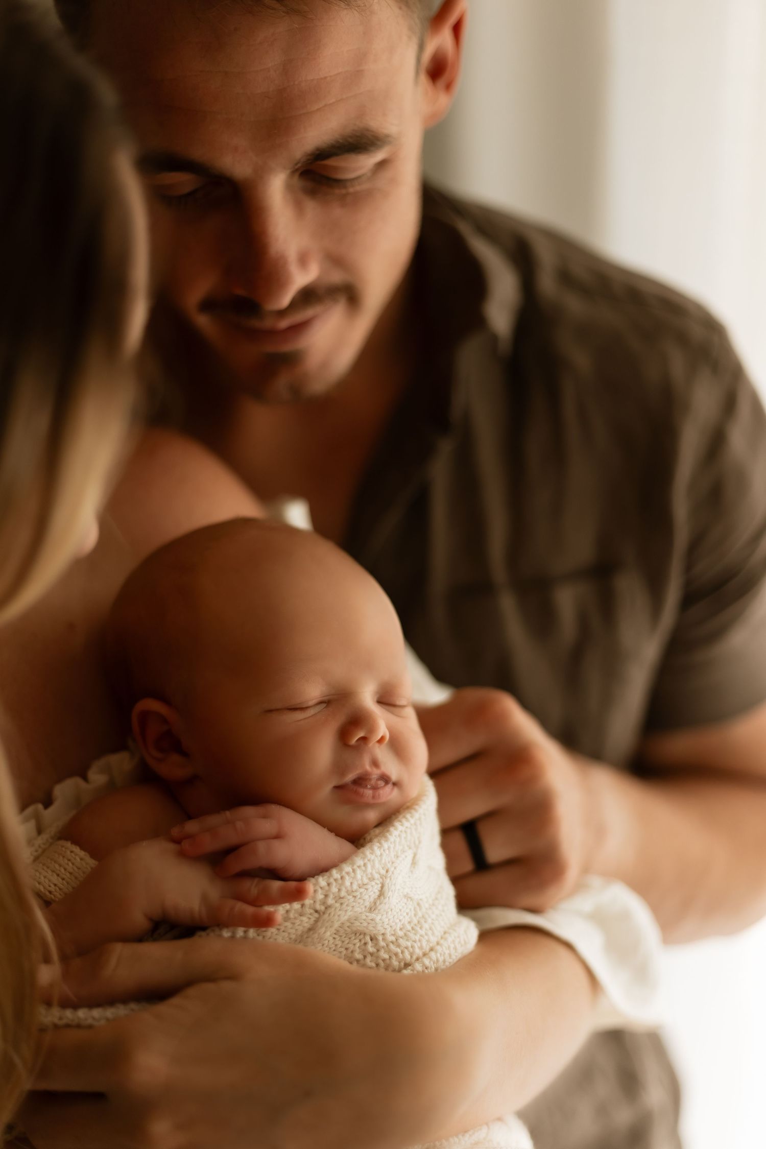 A sleeping newborn baby wrapped in soft white fabric is cradled tenderly while being admired in warm lighting.