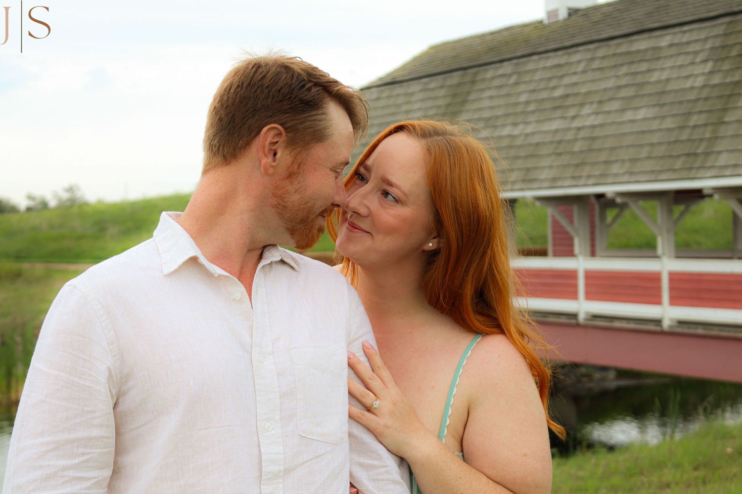 A couple in white and peach outfits embrace tenderly in front of a rustic barn setting during a romantic photoshoot.