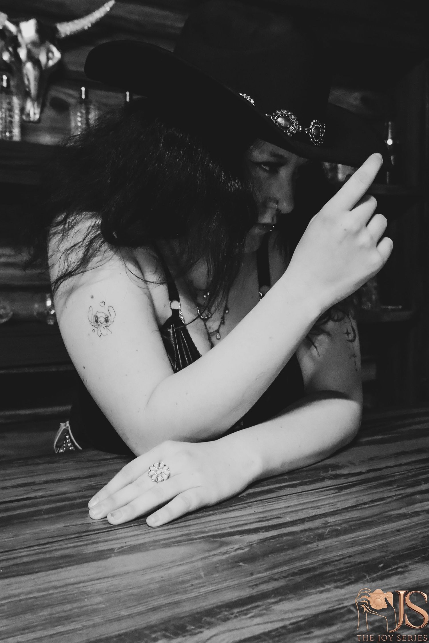 A moody black and white portrait at a wooden bar showing tattoos and a dark hat.