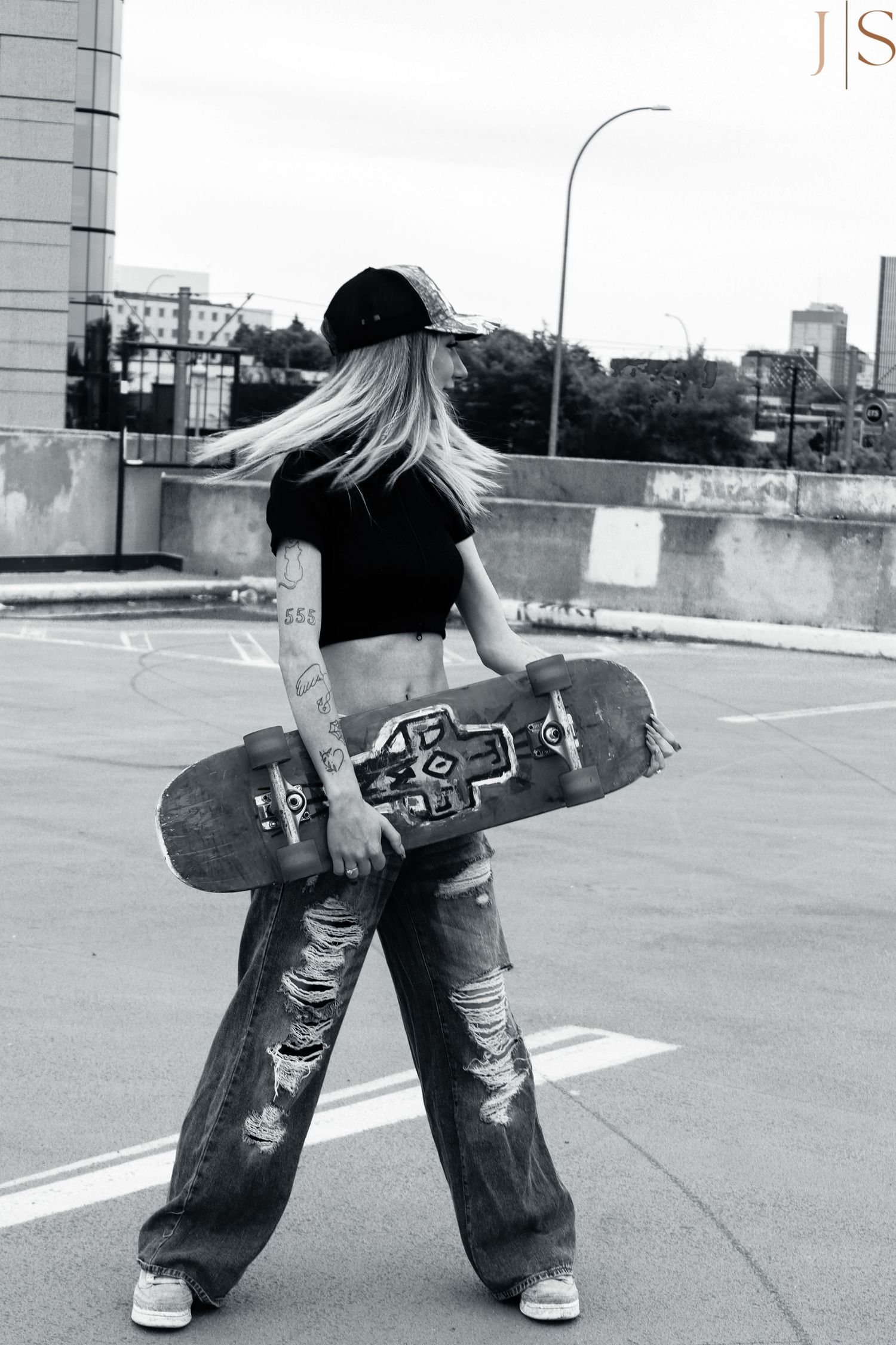 Black and white photo of a skater holding a skateboard in ripped jeans on an urban rooftop.