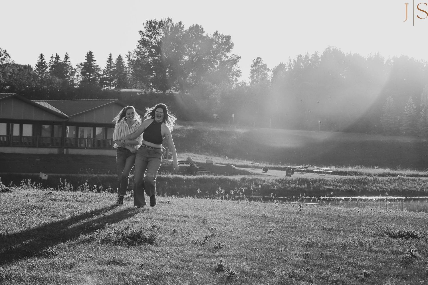 A couple stands together in a misty field at sunset near a rustic building in black and white photography.