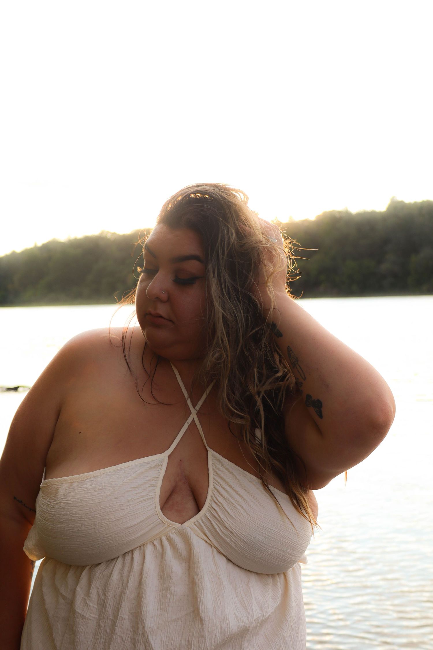 Person in white flowing dress poses at sunset by a serene lake in warm golden light.