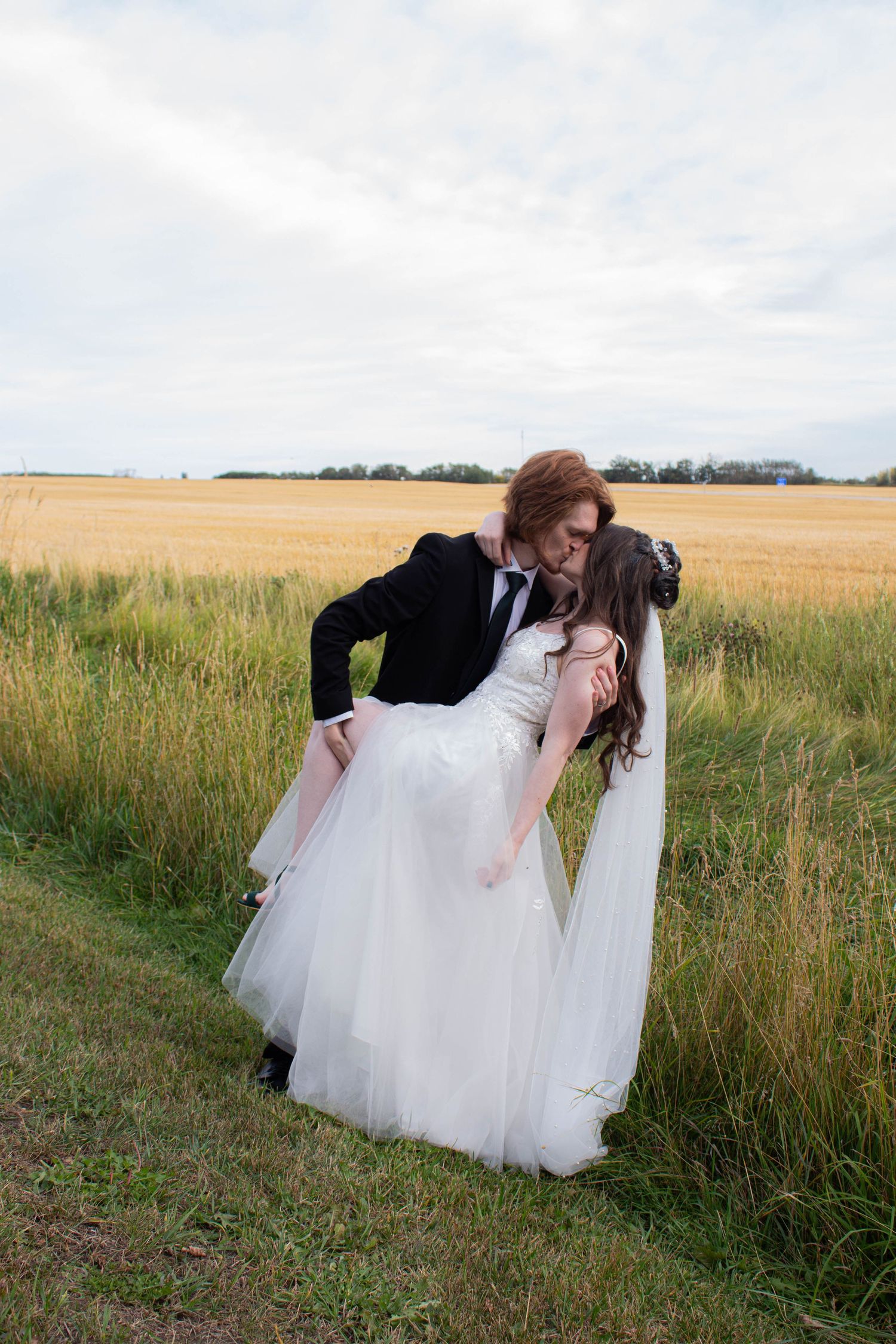 Romantic wedding dip pose in grassy field under cloudy sky.