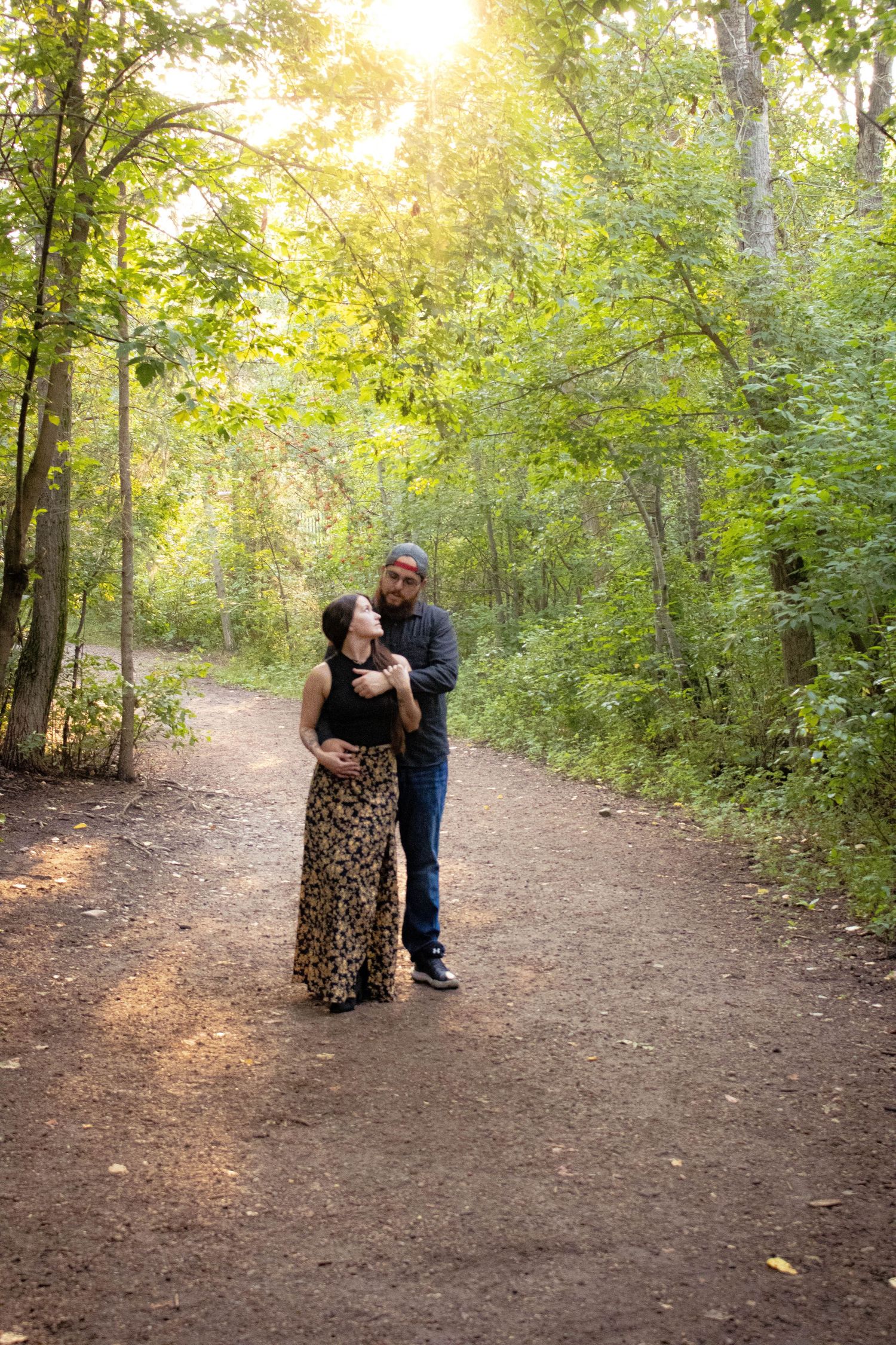 A couple sharing a tender moment on a sunlit forest path surrounded by green trees.