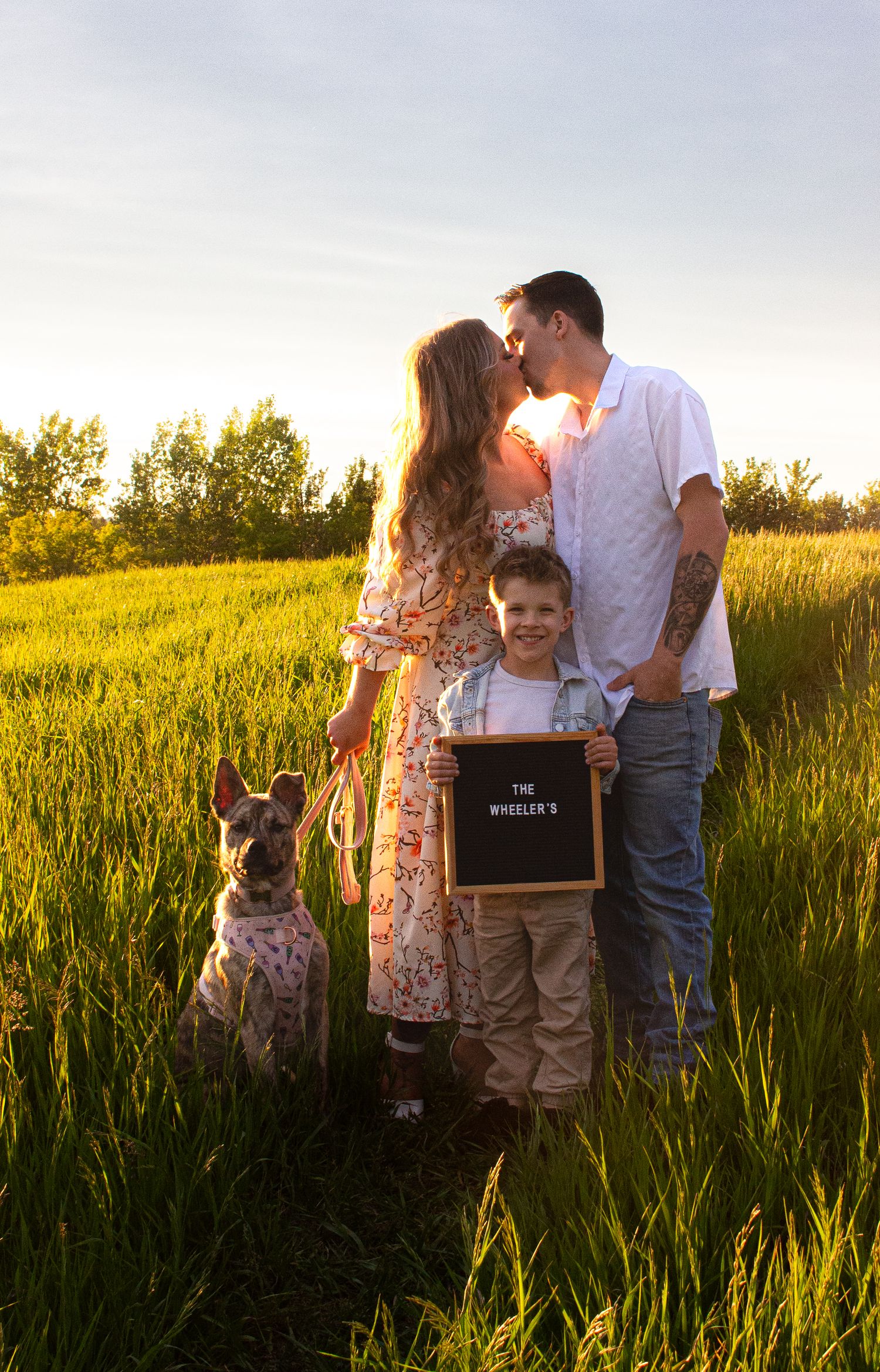 Family outdoor photoshoot at sunset with dog and chalkboard sign in grassy field.