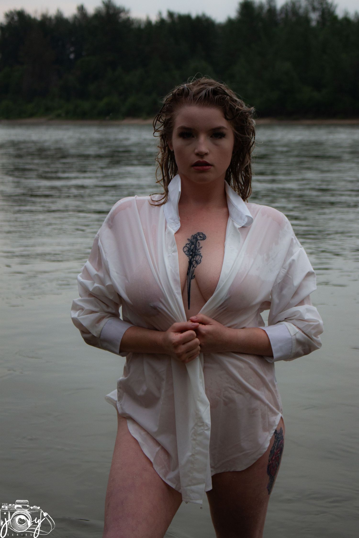 A figure in a wet white button-down shirt poses dramatically by a lake in moody outdoor photography.