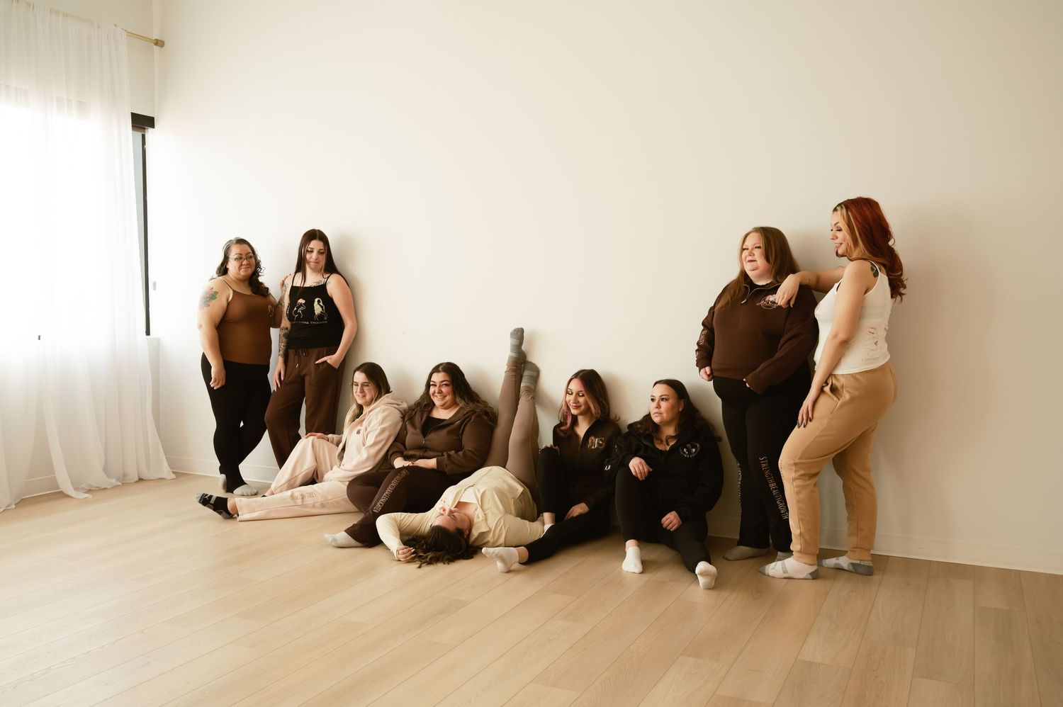 Group of eight women posing together in a bright minimalist studio, wearing coordinated brown and neutral tones.