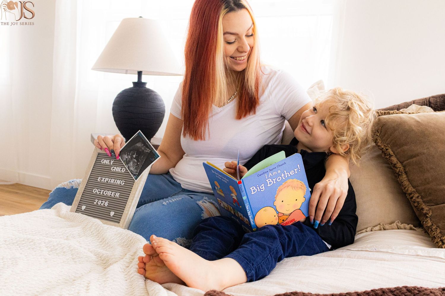 Mother and toddler reading Big Brother book together on bed with pregnancy announcement chalkboard sign.