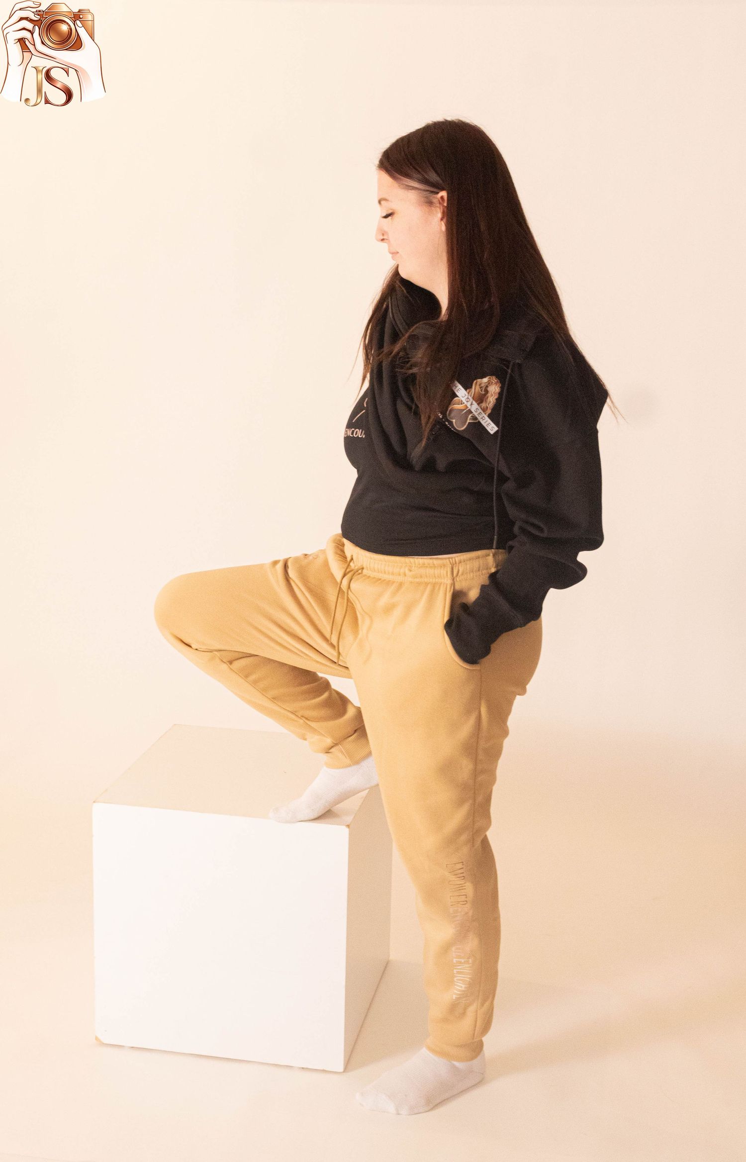 Person in black hoodie and beige pants leans forward over white box against light pink background.