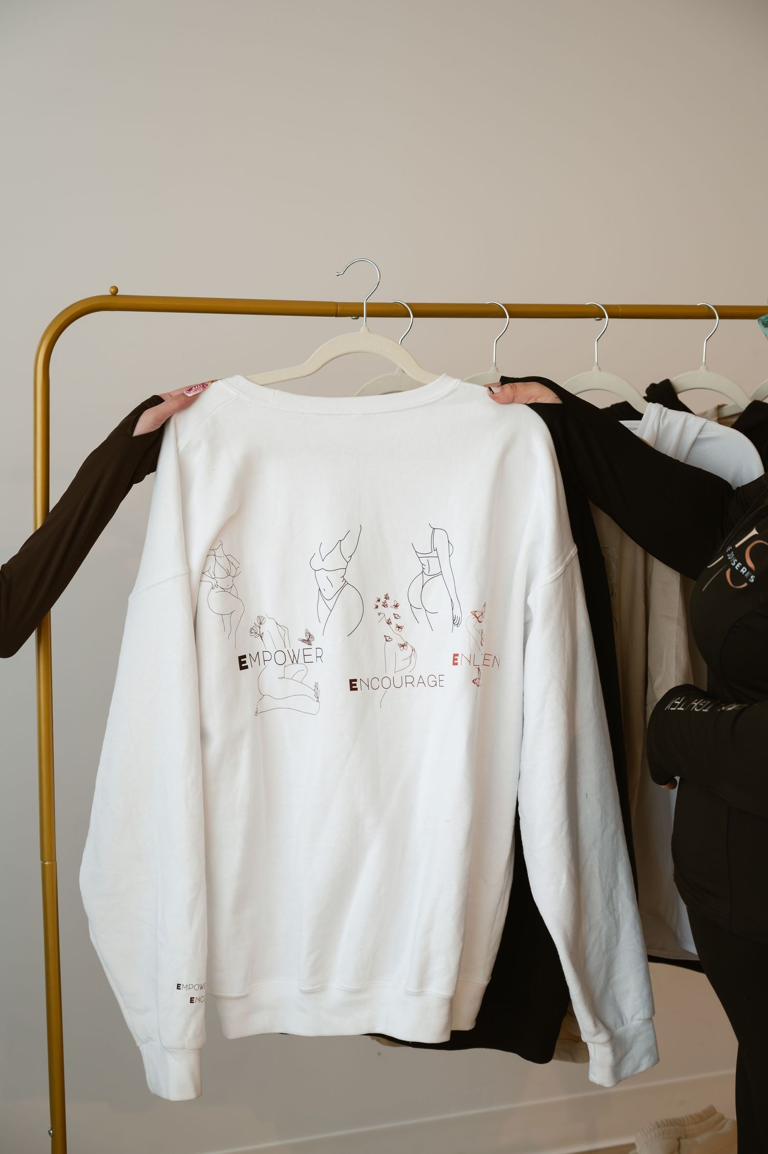 White sweatshirt with Empower, Enlighten, Encourage graphic print hanging on a gold clothing rack.