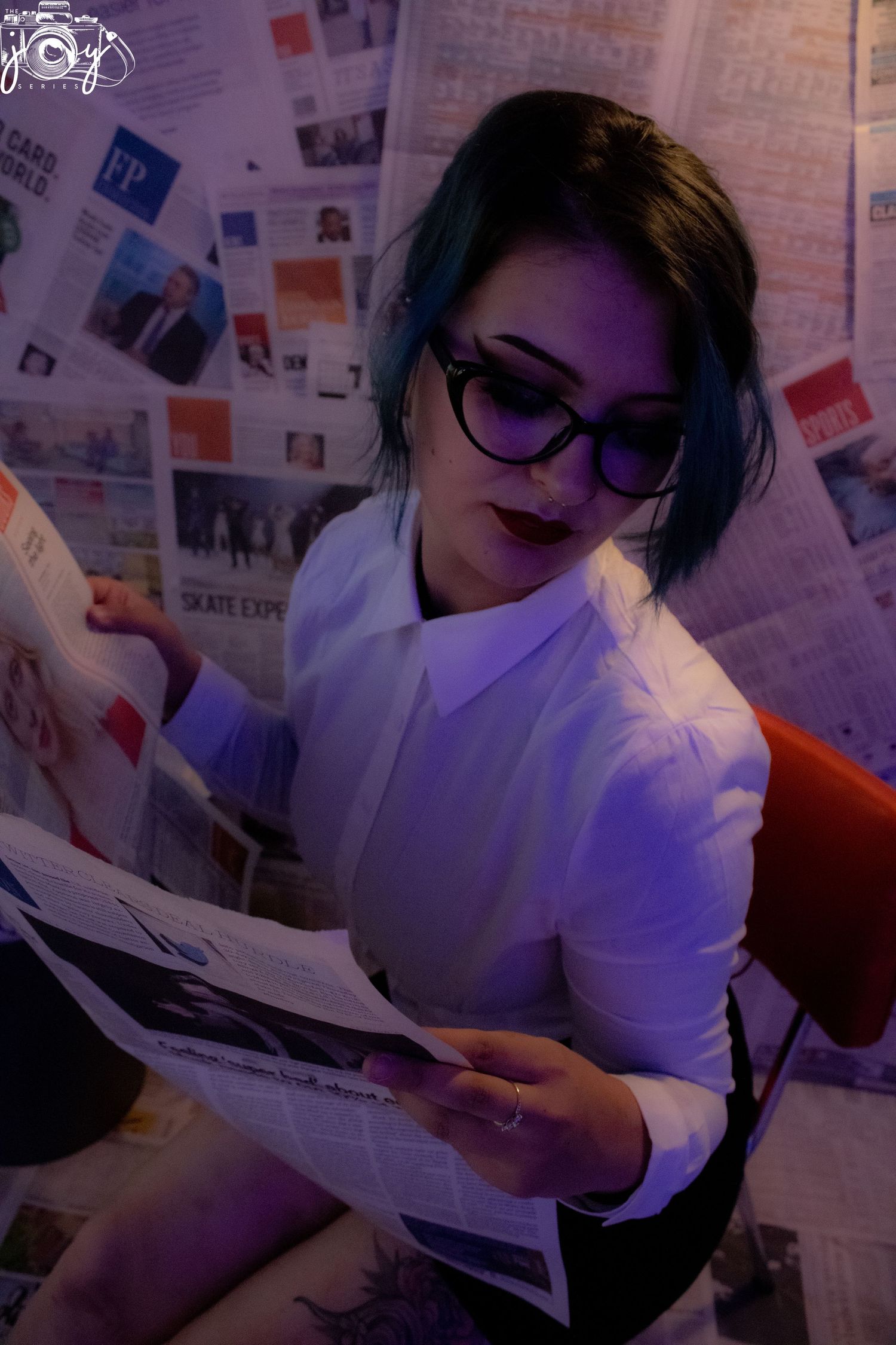 A person in glasses and white shirt reads papers against a wall covered in photos and articles.