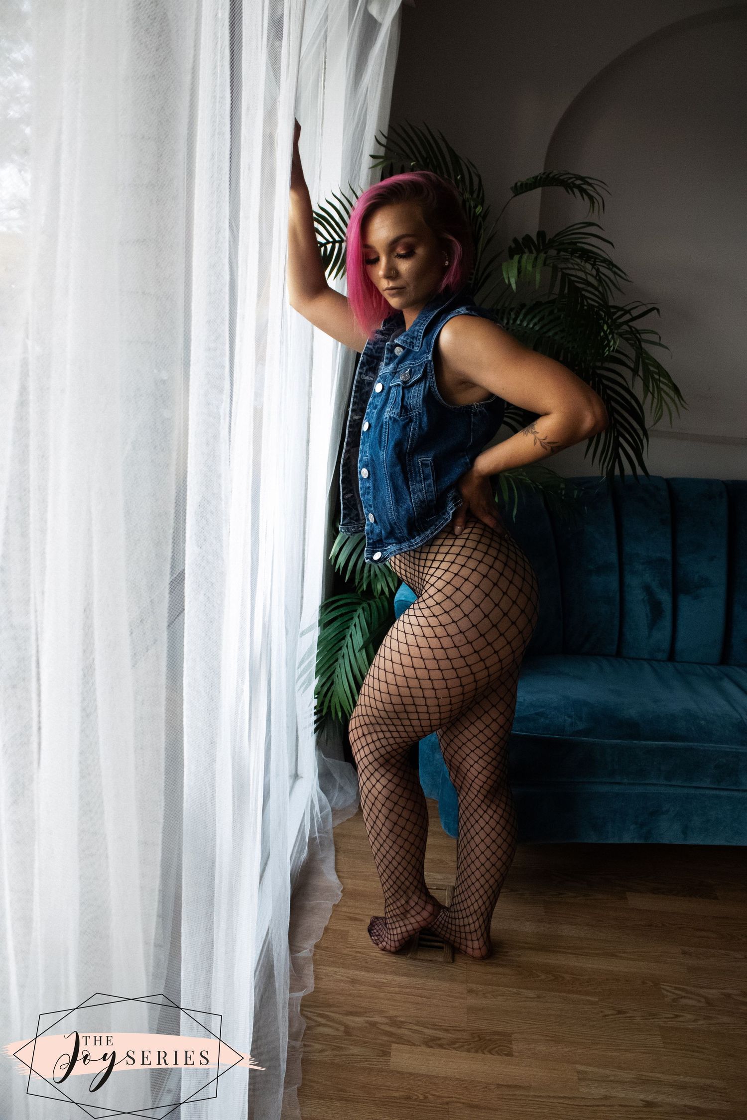 Person in denim vest and fishnet stockings poses by window against blue velvet couch and tropical plants.