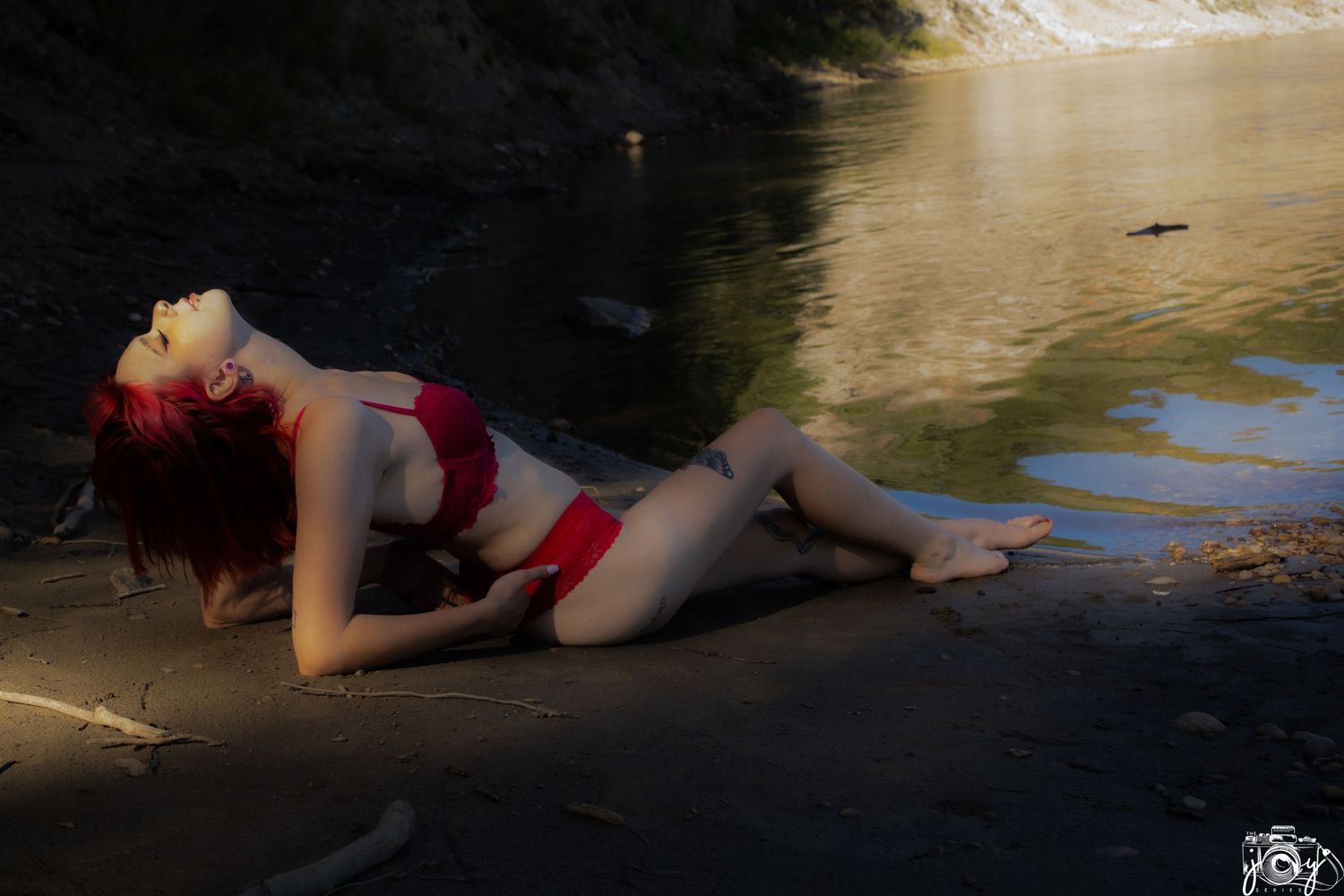 A figure in a red swimsuit reclines on a rocky riverside in dramatic sunlit lighting.