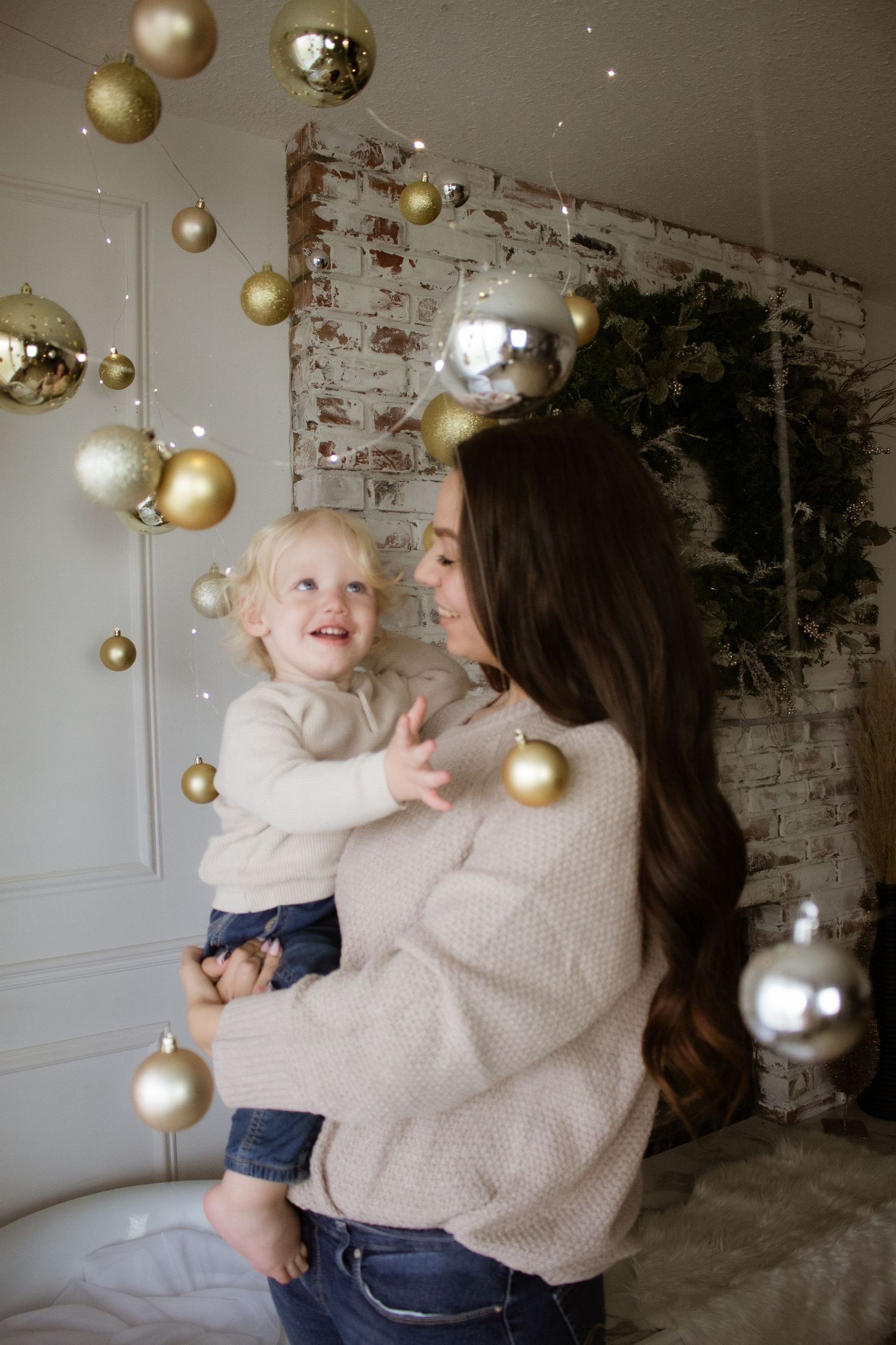 Holiday scene with floating gold ornaments and person holding toddler against white brick wall.