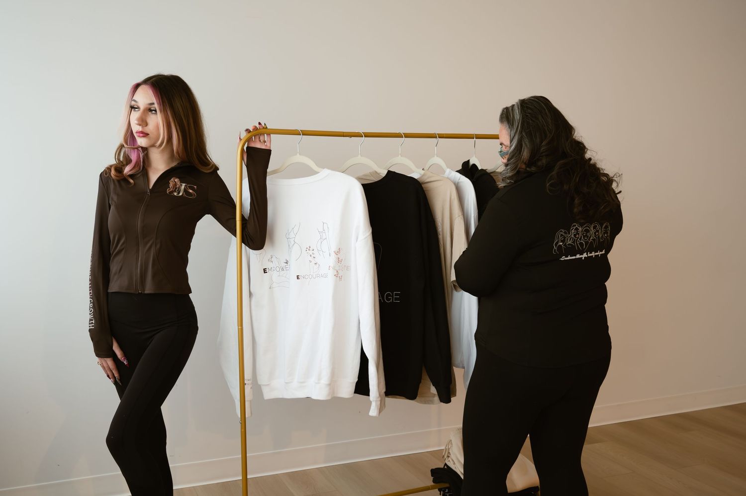 Two women arrange clothing on a gold rack displaying white, black, and brown garments against a white wall.