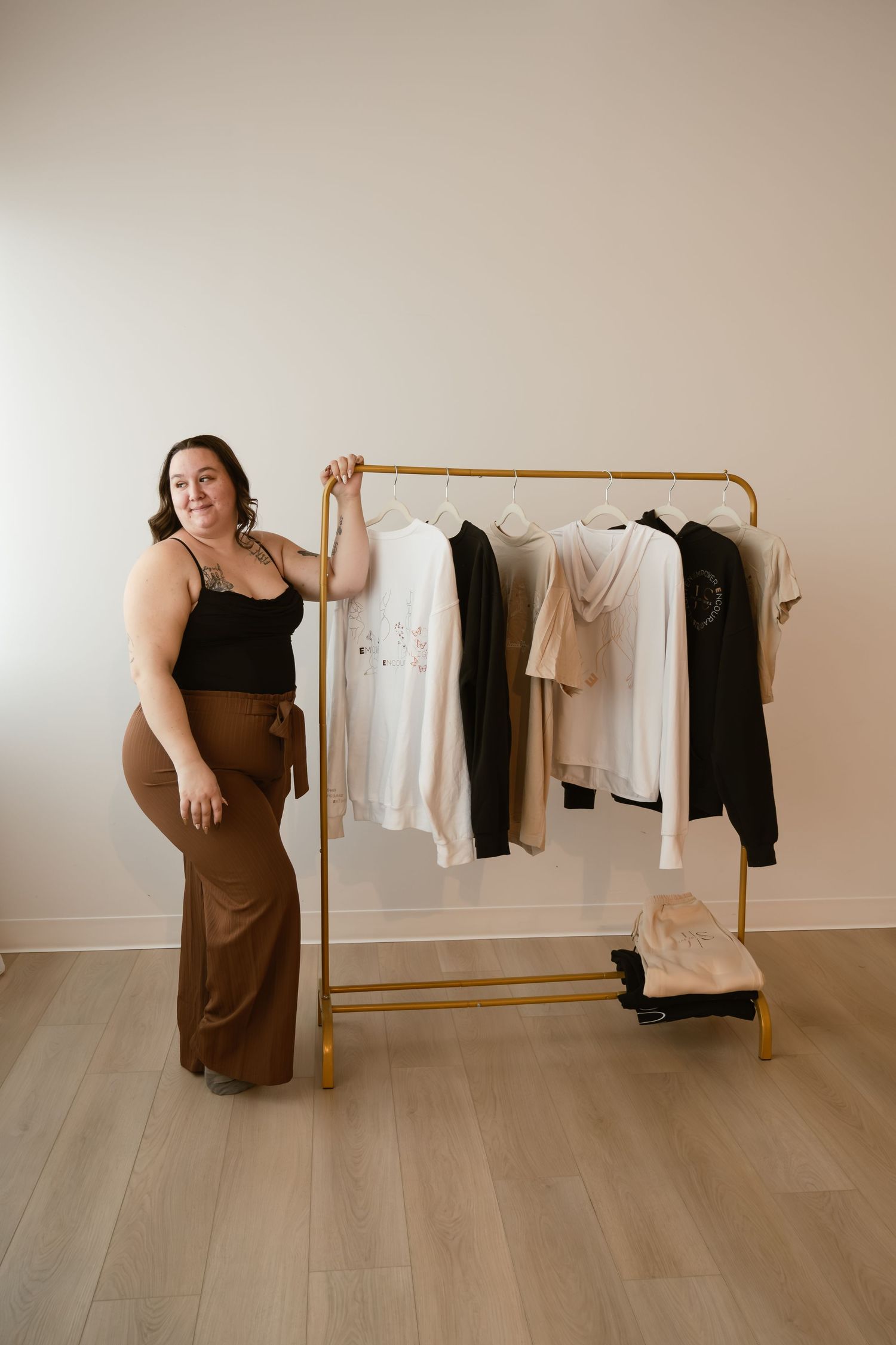 Woman in black top and brown pants posing beside a gold clothing rack with neutral-toned garments.