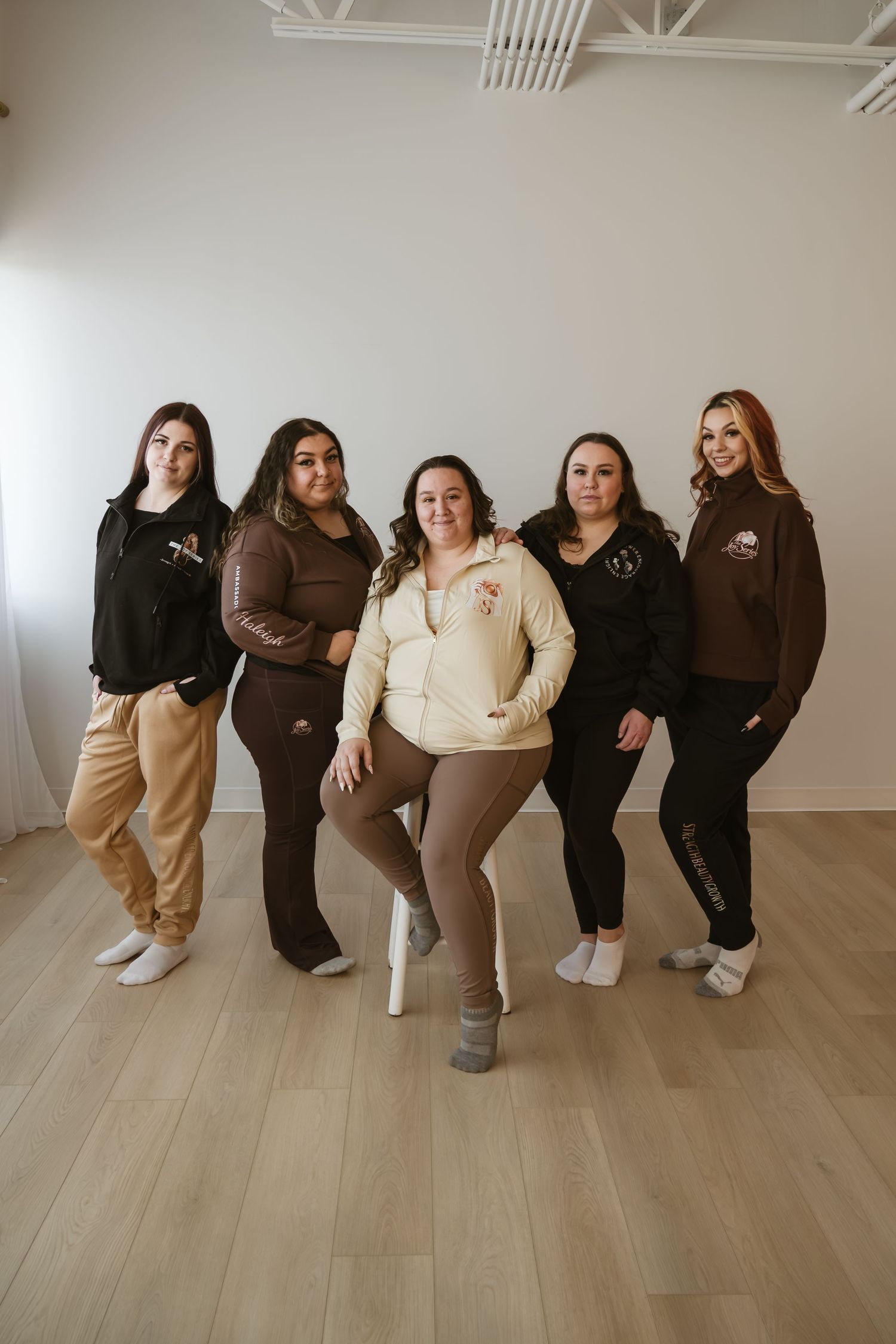 Five women posing together in brown and black branded loungewear in a bright studio with wood floors.