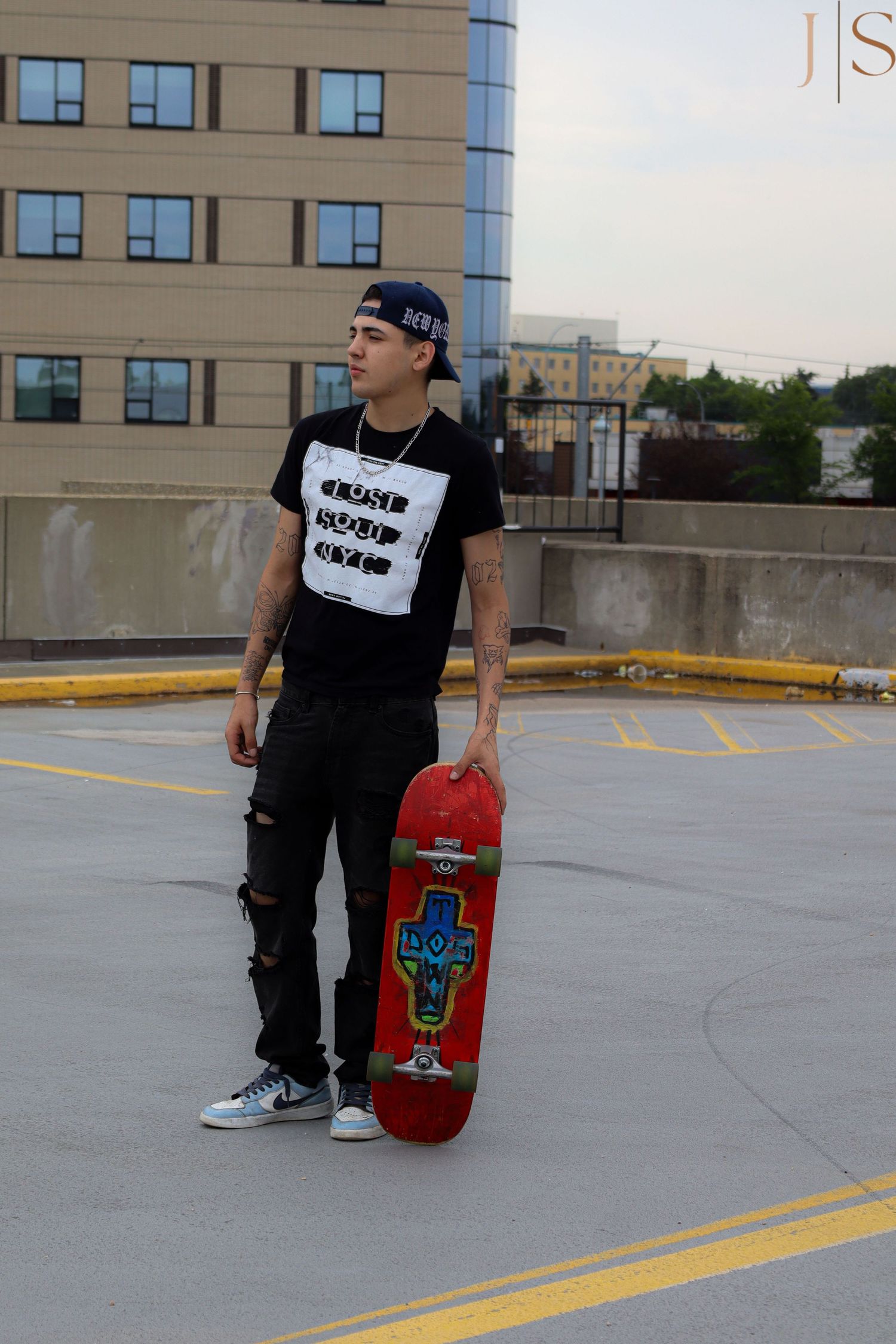 A skateboarder in a black t-shirt and ripped jeans holds a red skateboard in an urban parking lot.