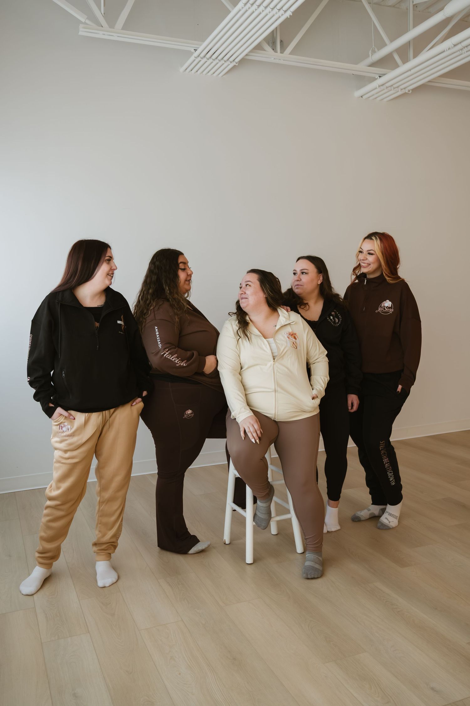 Five women in matching brown and neutral loungewear pose together in a bright minimalist studio space.