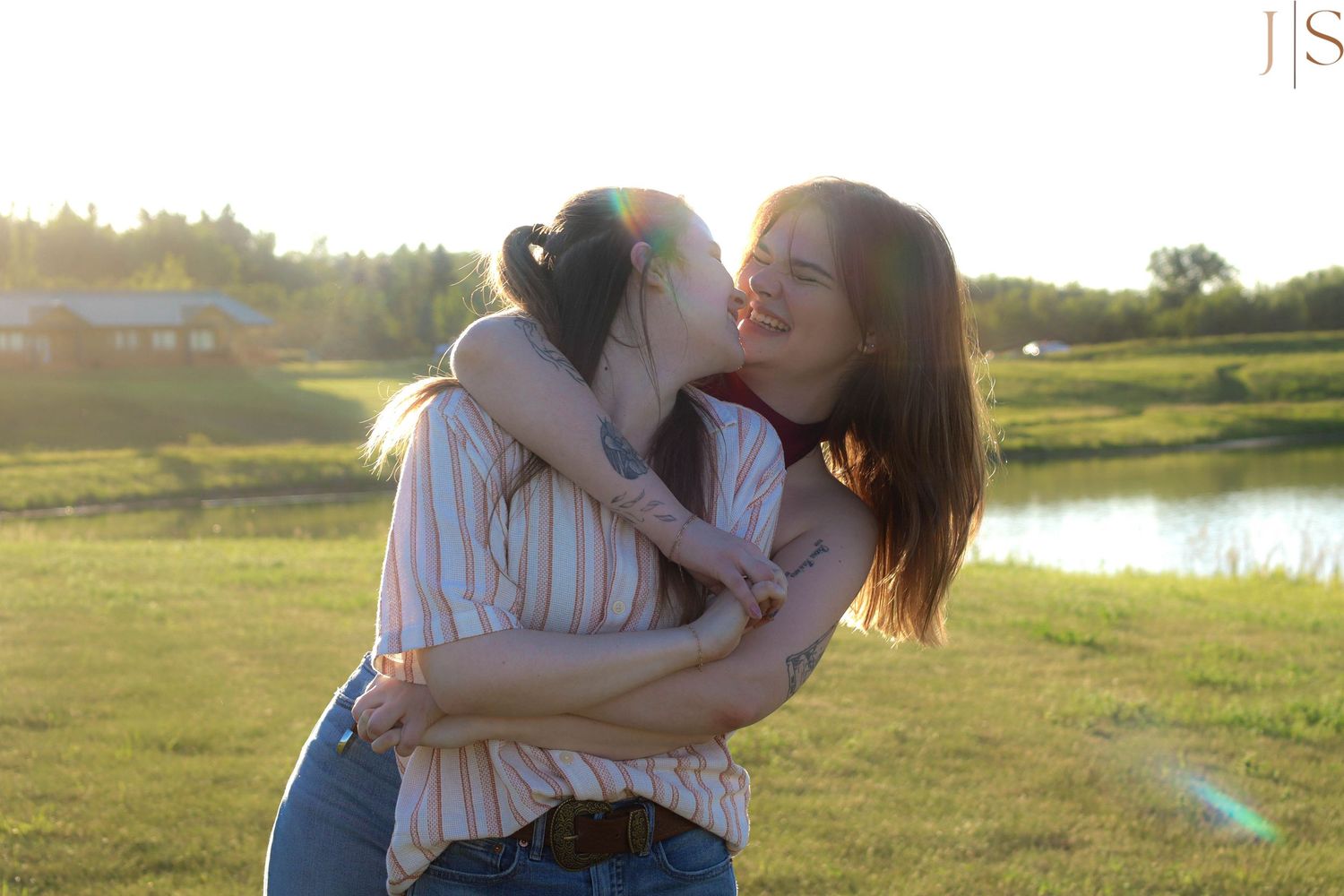 Two people embracing joyfully in a sunlit field by a pond during golden hour.