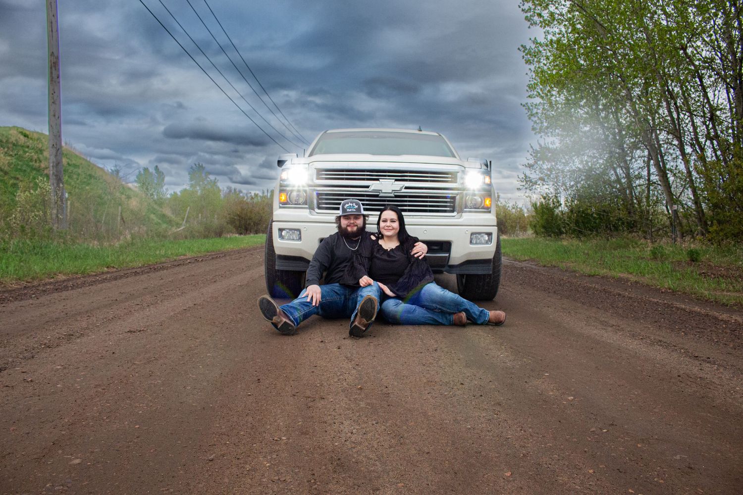 A couple poses sitting on a dirt road in front of a white Chevrolet truck during overcast weather.