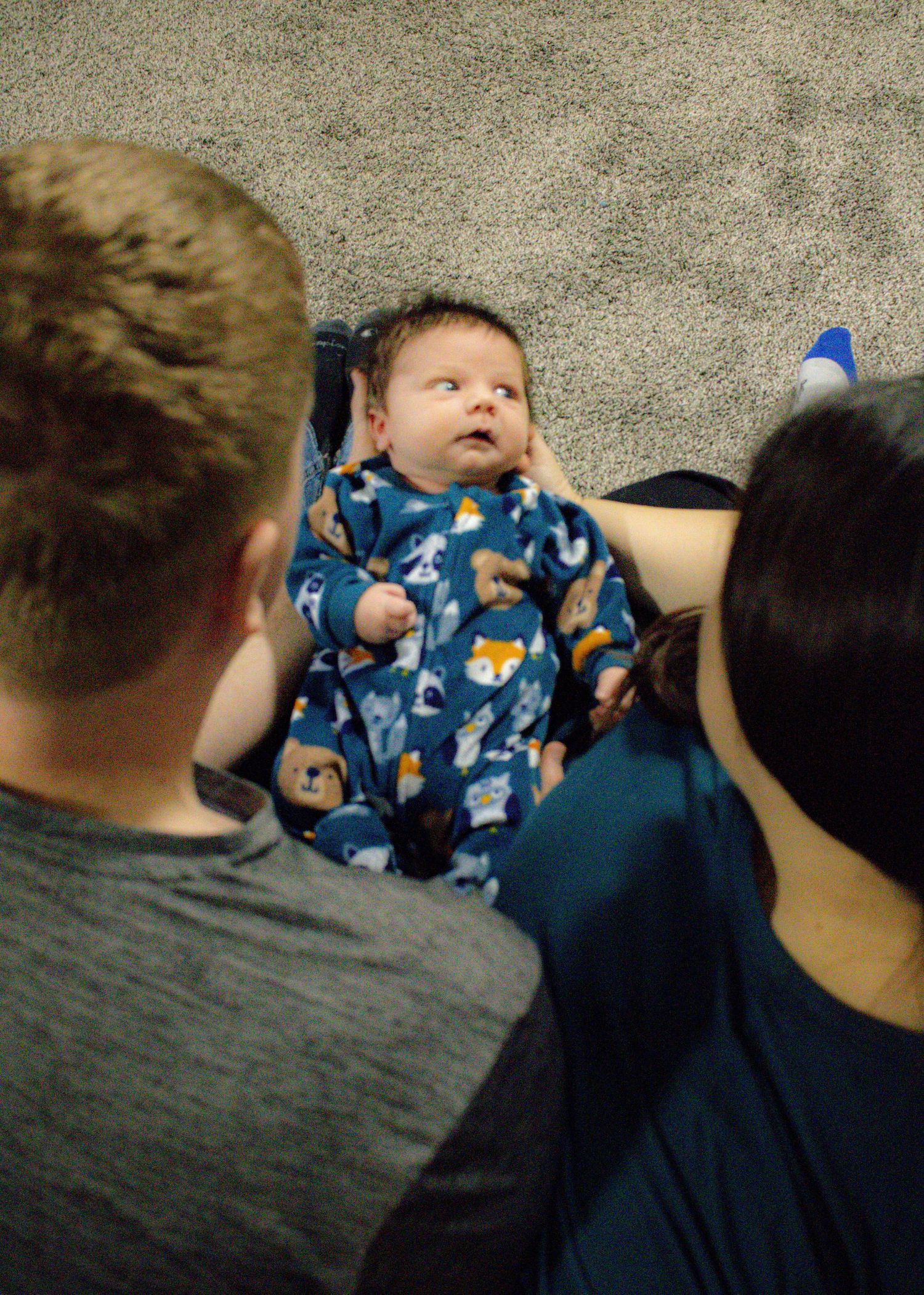 Baby wearing blue patterned pajamas lying on carpet surrounded by family members.