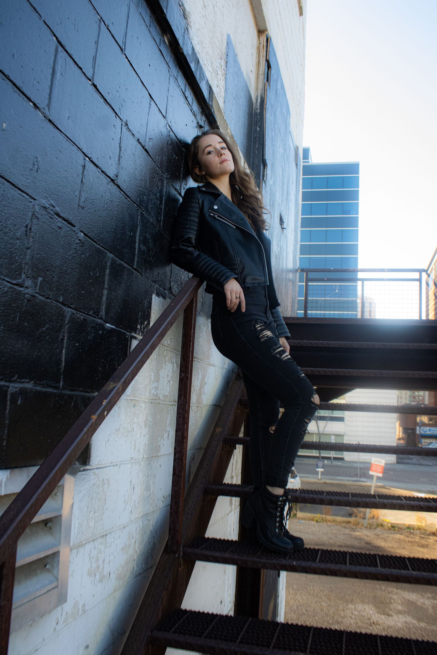 Model wearing all black poses on metal stairs with a modern building in the background.