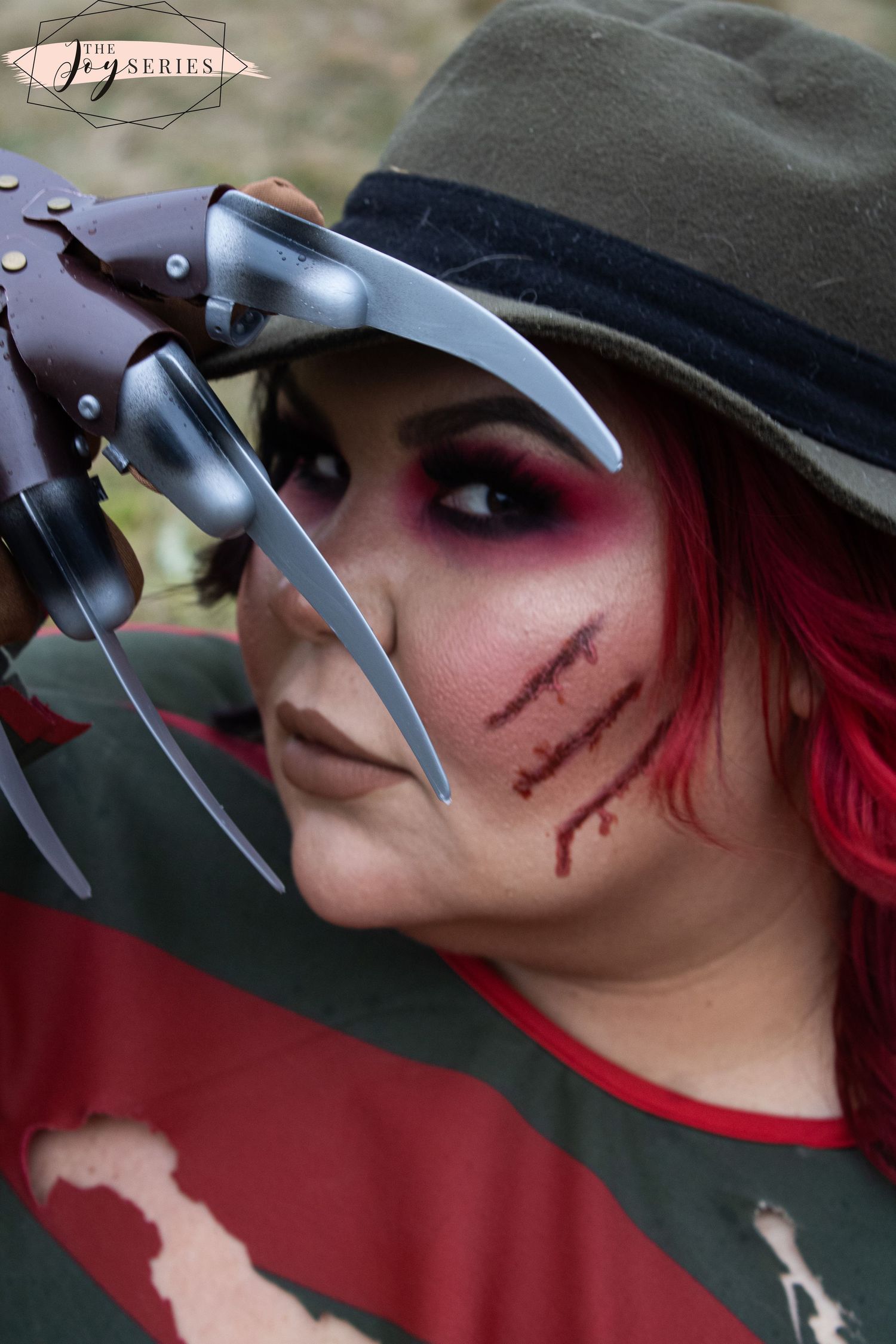 A horror-themed cosplay featuring dark makeup, scars, a brown fedora, and metallic claws against a red background.