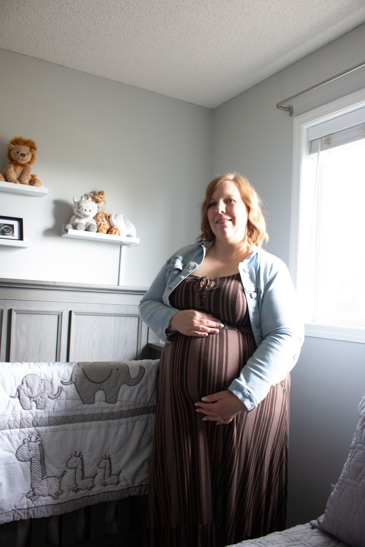Expectant mother in brown dress and blue cardigan poses in nursery with stuffed animals on shelves.
