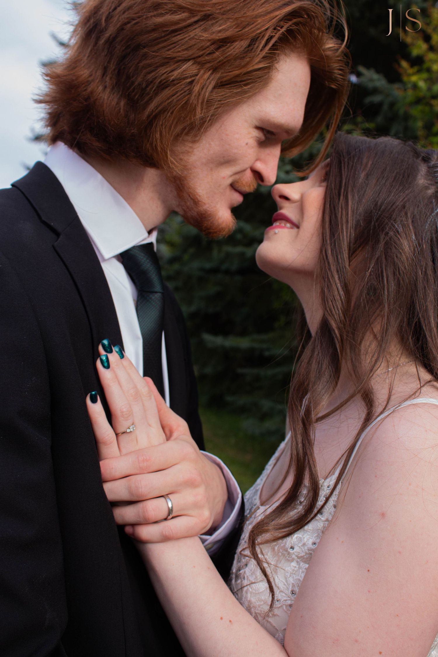 A romantic couple in wedding attire share an intimate moment outdoors, with the bride wearing a sparkly dress and showing her ring.