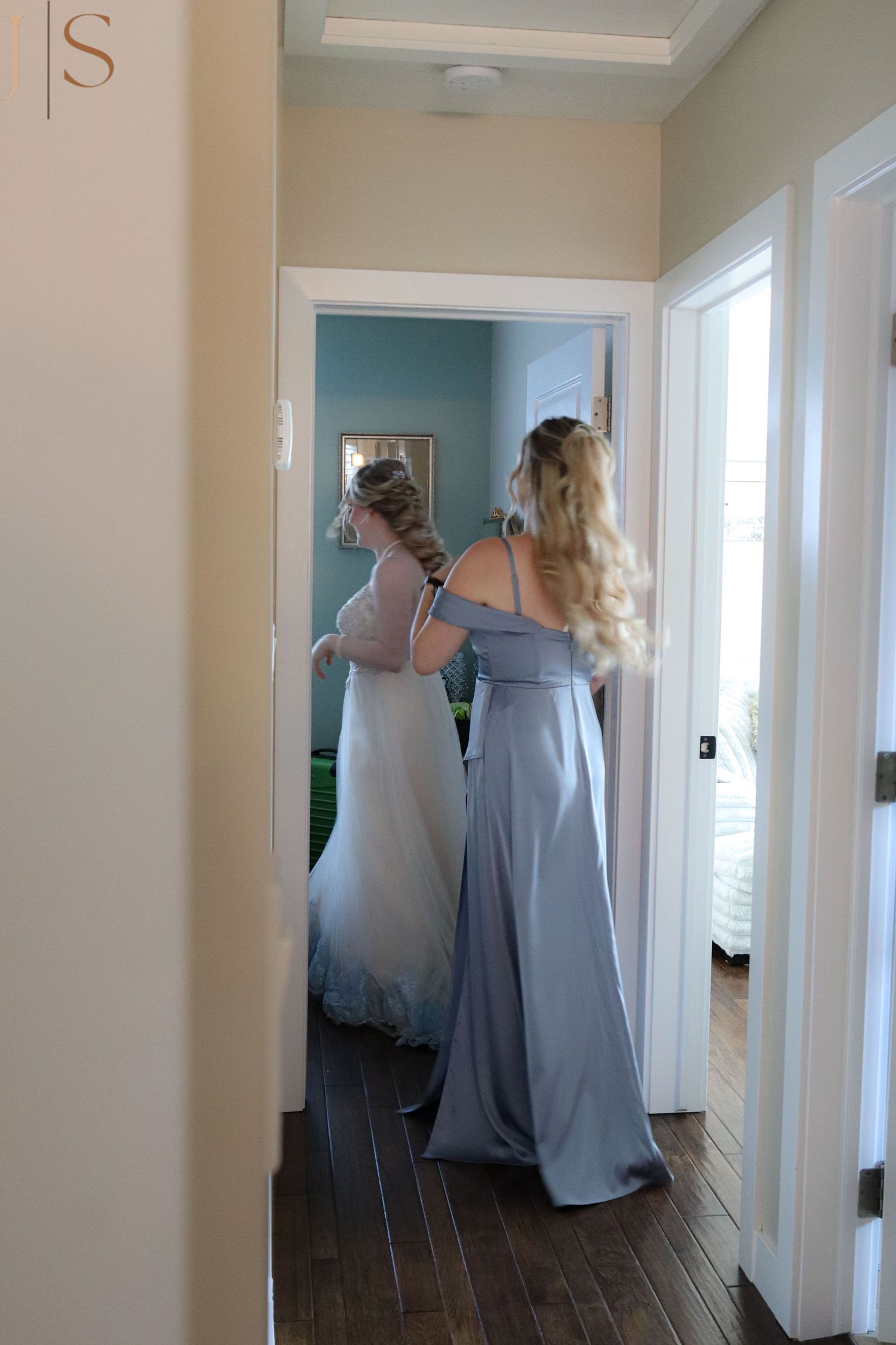 Two women in formal dresses help with wedding preparations in a hallway with light blue walls and hardwood floors.