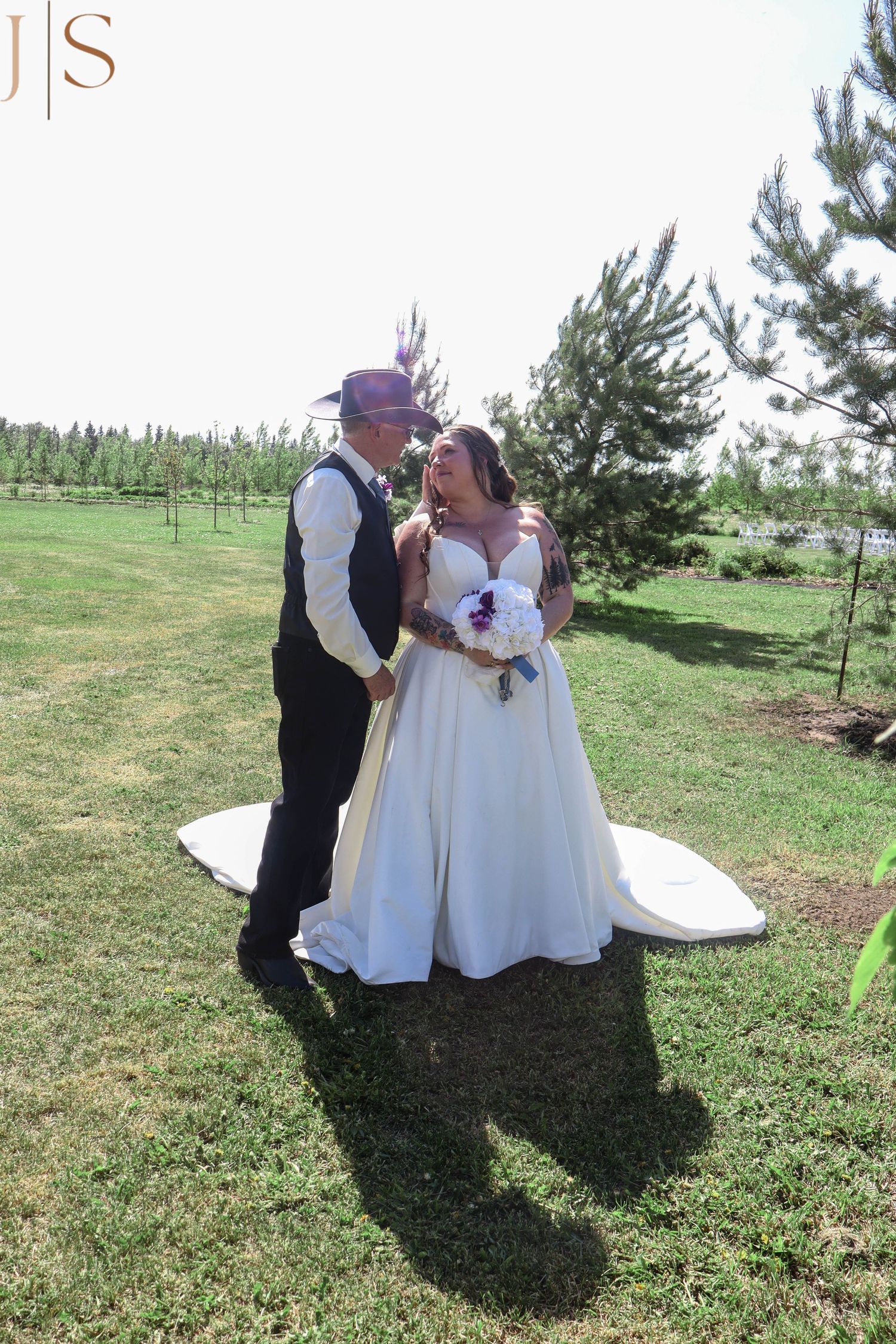 A couple shares a tender moment outdoors during a wedding celebration on a sunny day at a tree-lined venue.