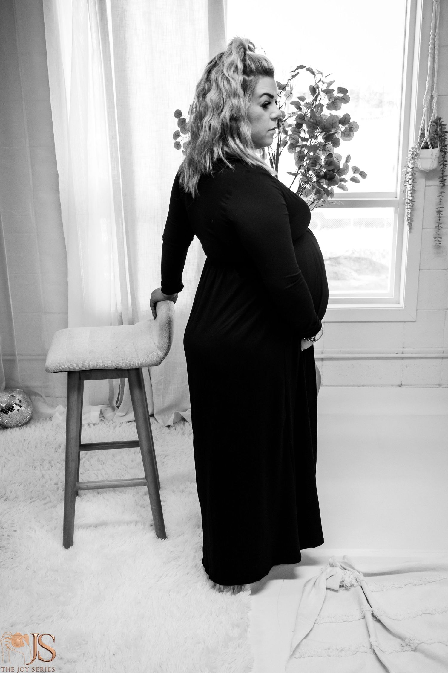 A black and white maternity photo shows a woman in a long black dress posing next to a wooden stool near a window.