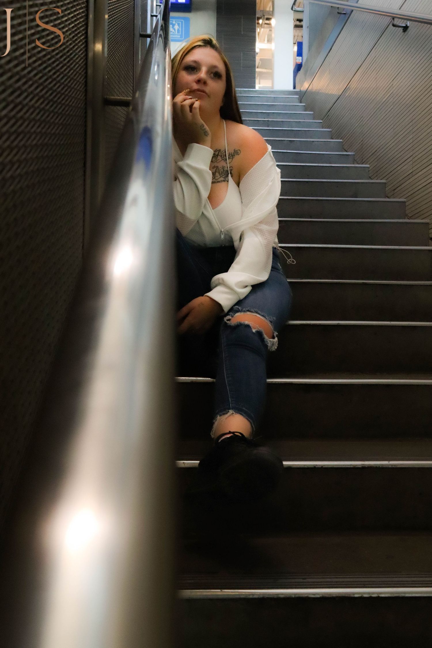 A person sits on dark stairs with a handrail while wearing a white sweater and jeans in a dimly lit stairwell.