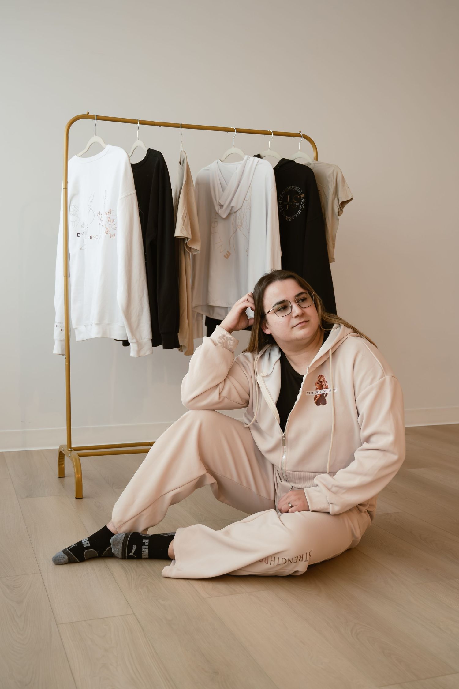 Woman in pink tracksuit sits on floor near gold clothing rack with various garments in minimalist white room.