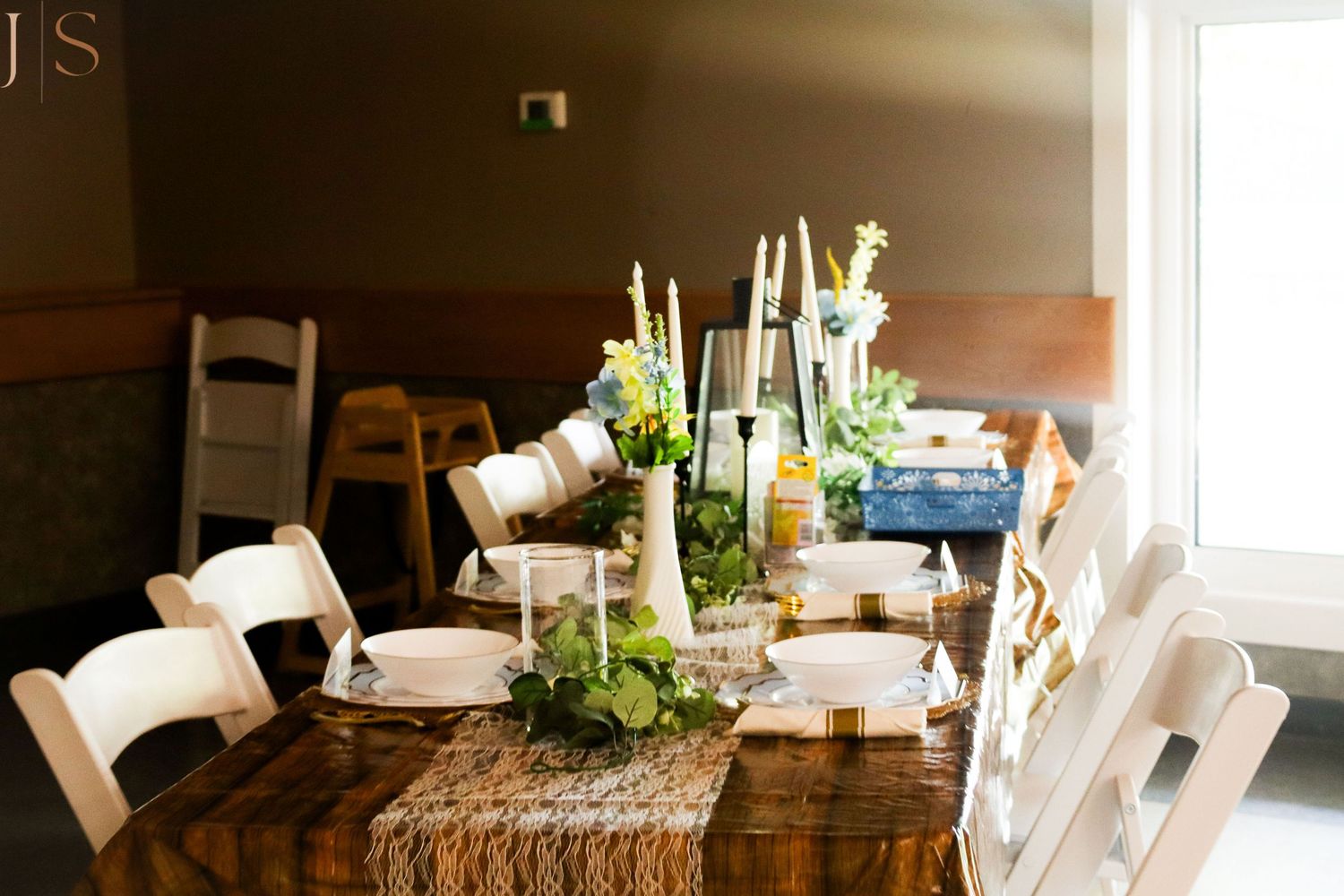 Rustic dining table set with white chairs, burlap runner, and elegant candle centerpieces in a warm-lit room.