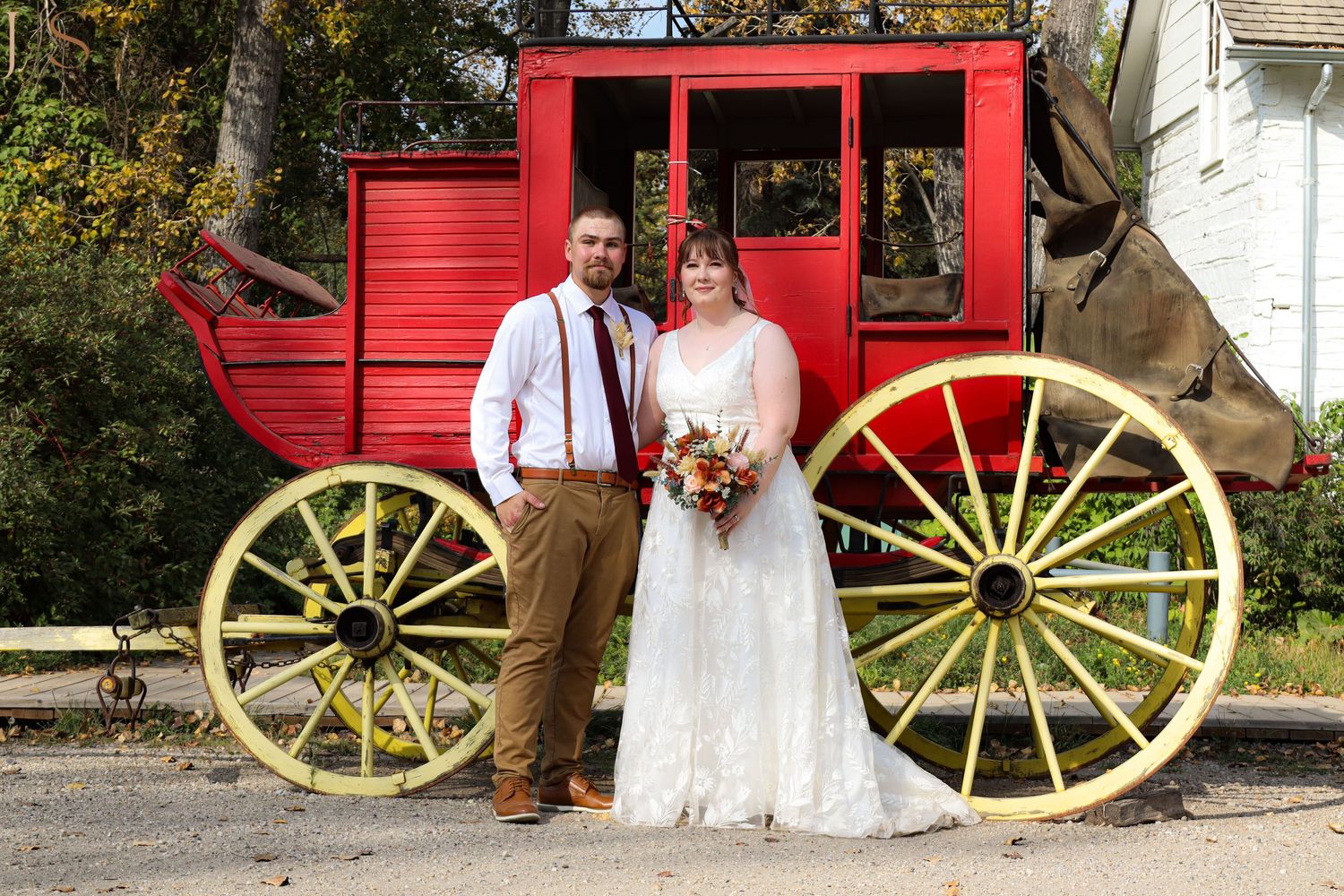 Couple poses in front of a vintage red carriage with large yellow wagon wheels for rustic wedding photos.