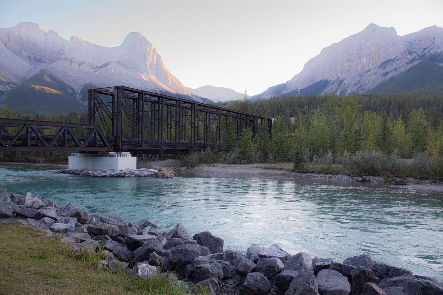 Historic iron bridge crosses mountain river with rocky shore and dramatic peaks in background.