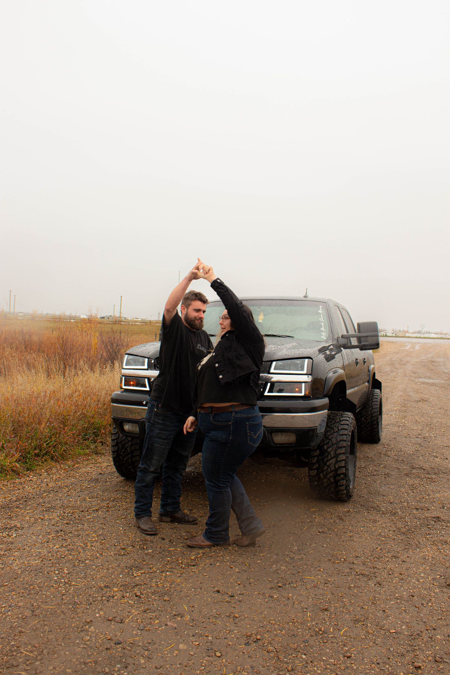 Couple making heart shape with arms in front of lifted truck on dirt road.