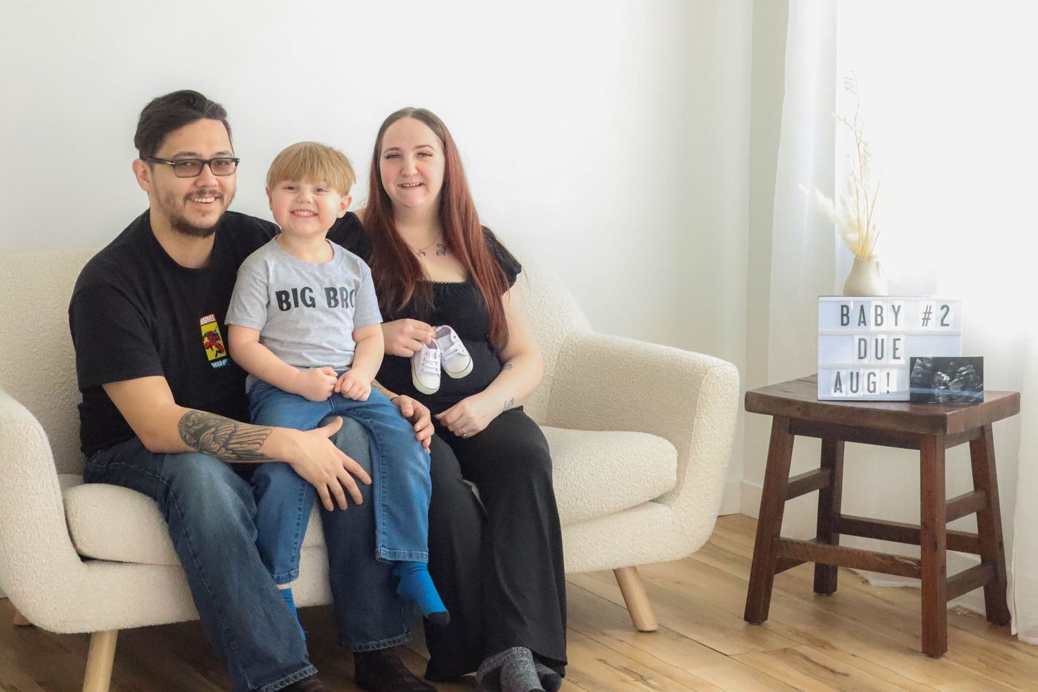 Family portrait in white room with pregnancy announcement props on side table.