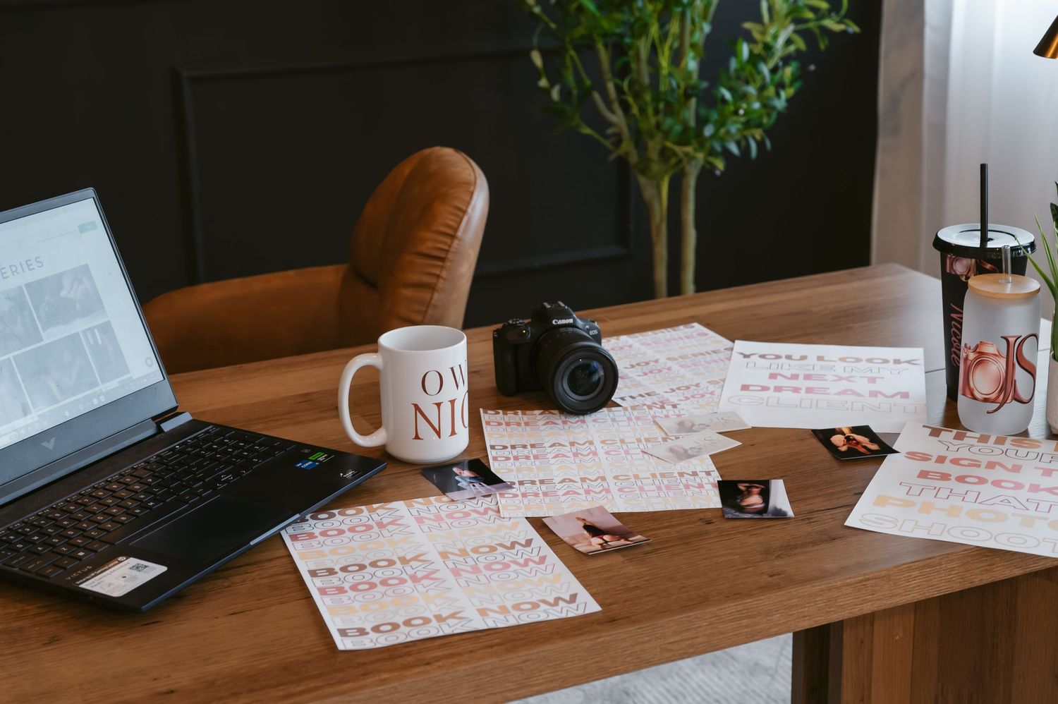 Wooden desk with laptop, camera, OWN IT mug, planner sheets, and a drink cup in a cozy home office setup.