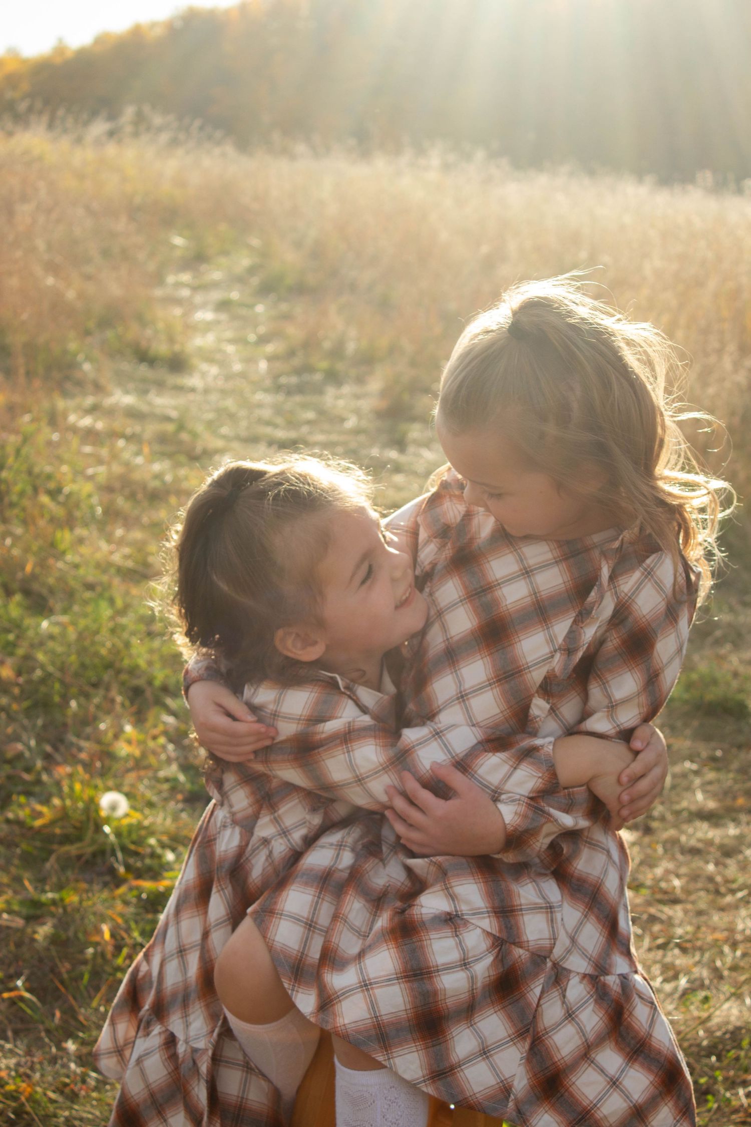 Two children in matching plaid outfits embrace and laugh together in a sunlit meadow.