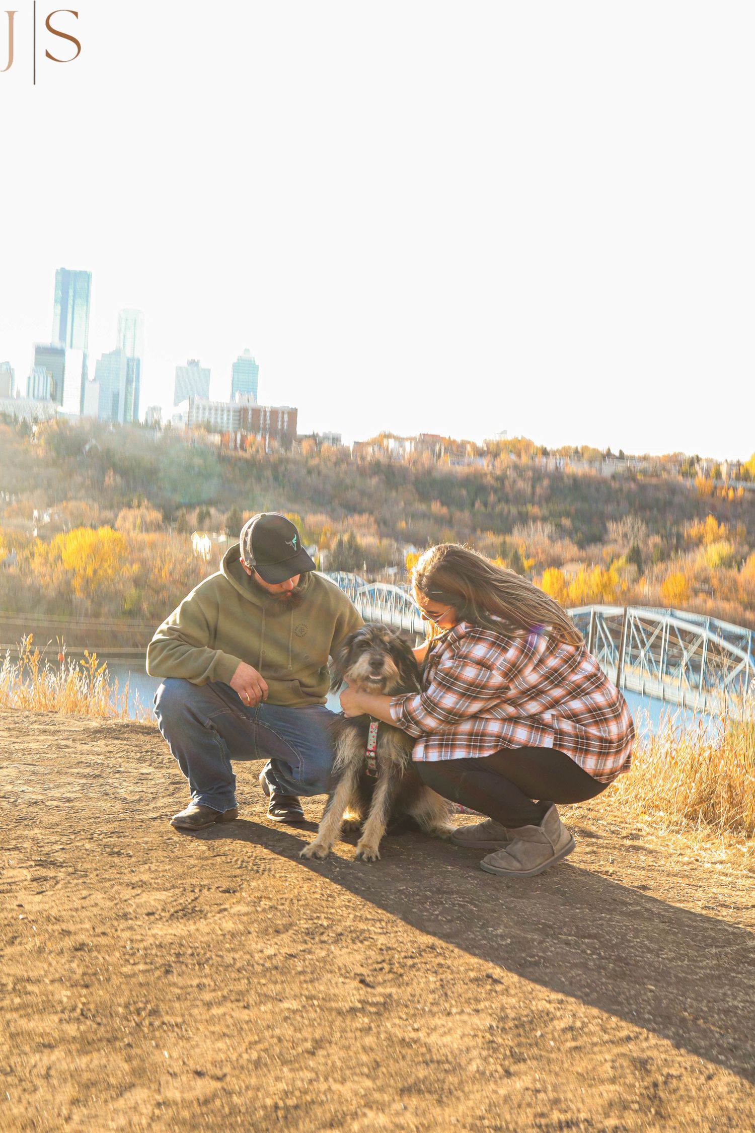 Couple crouching with their dog on a dirt path during autumn with city skyline and fall foliage in background.