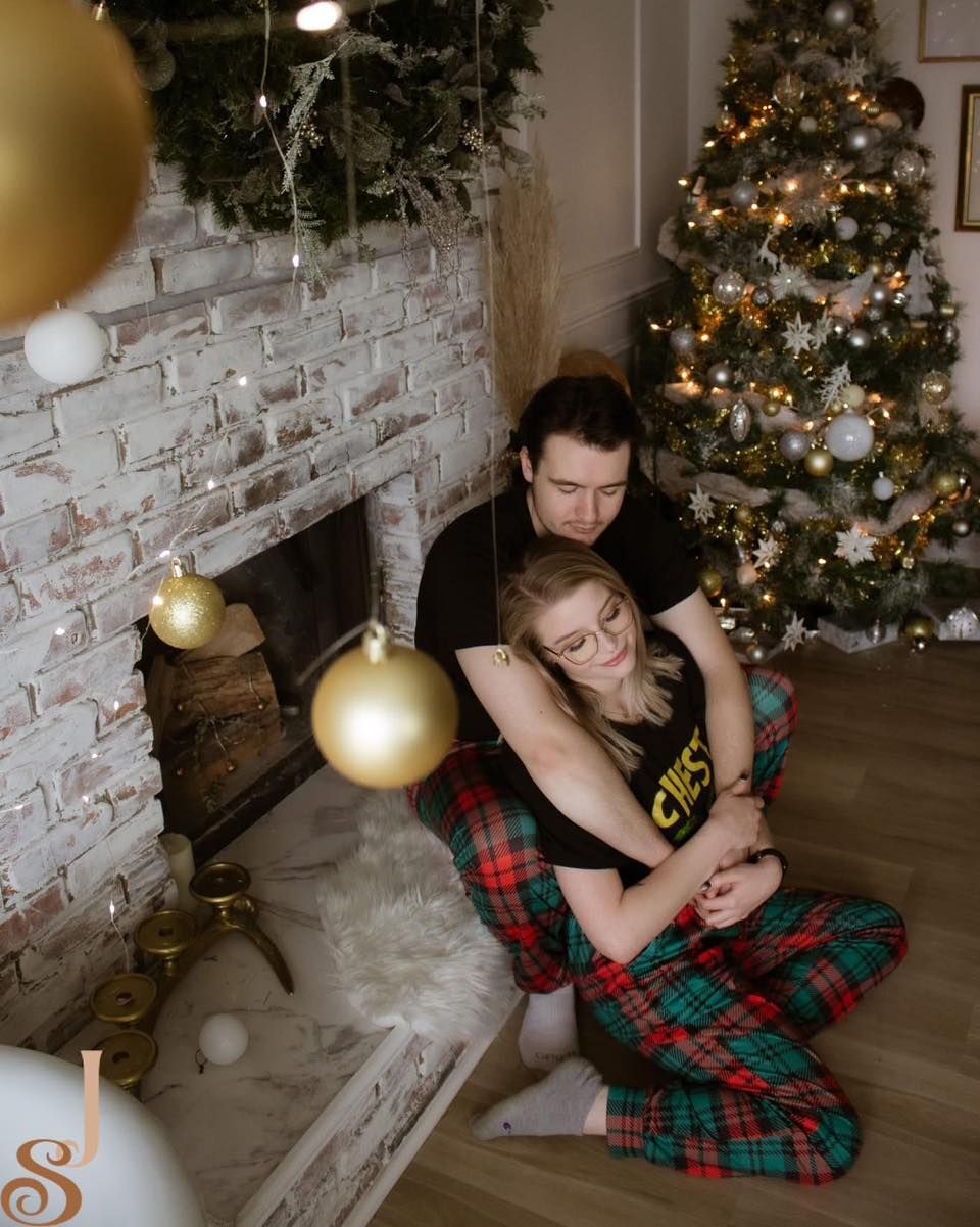 A couple in matching plaid pajamas share tender moments near a decorated Christmas tree and white brick wall.
