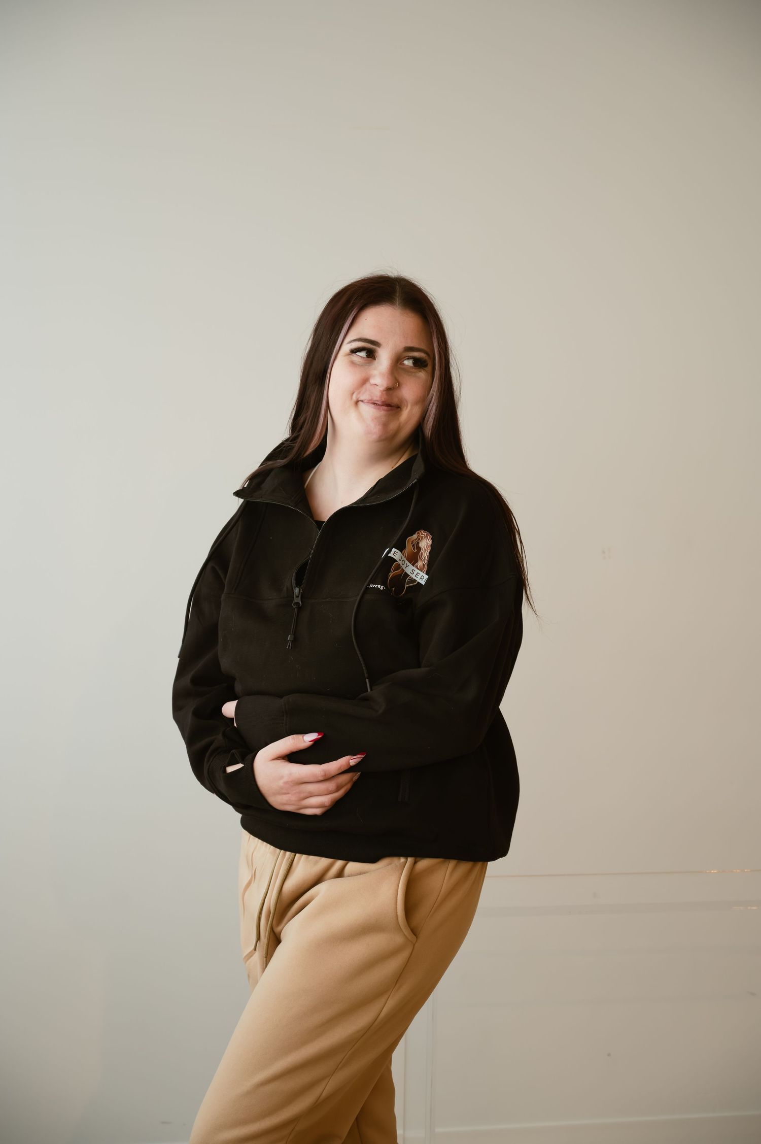 A young woman in a black quarter-zip pullover and tan pants poses smiling against a light gray background.